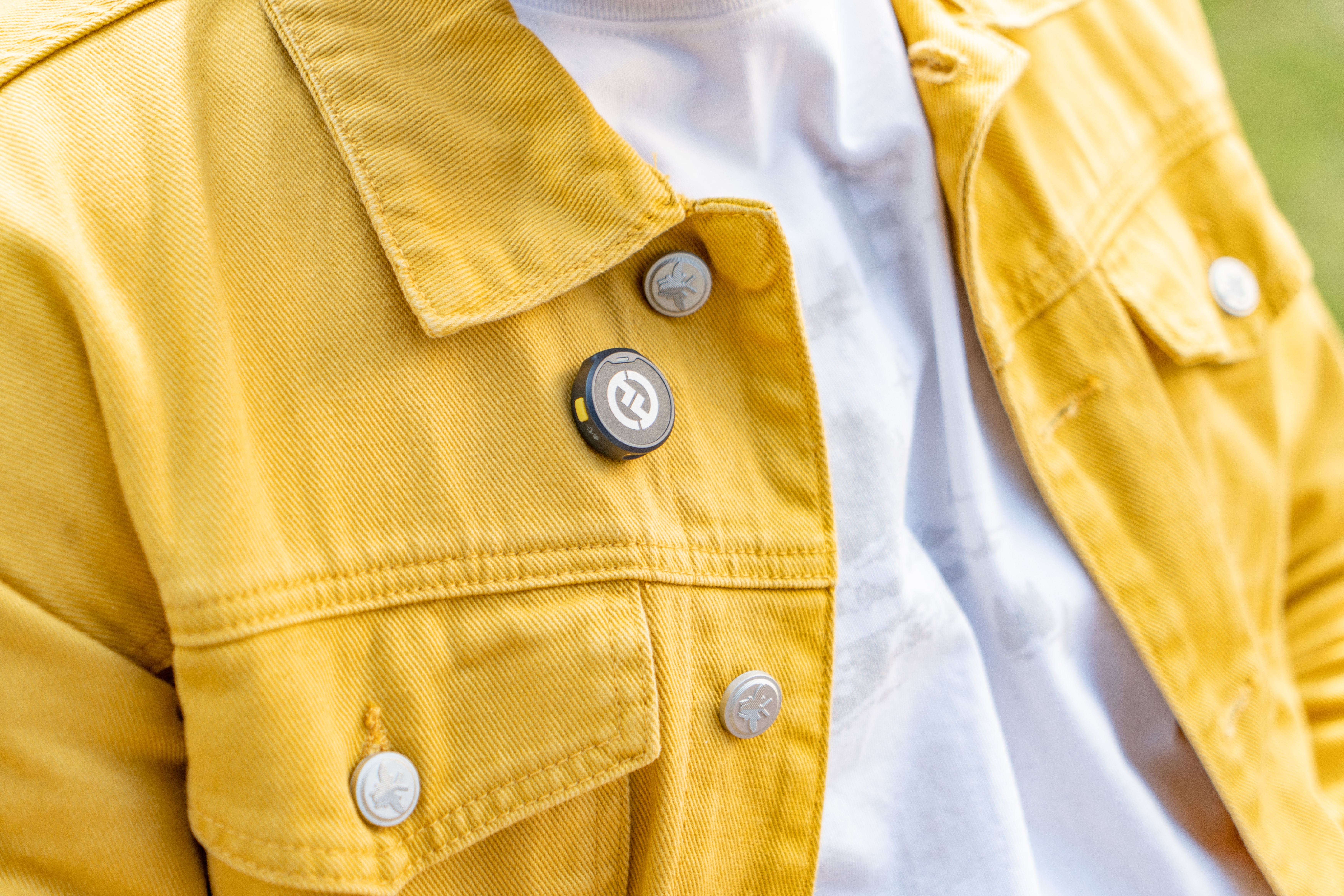 Osoba nosząca żółtą kurtkę dżinsową z przypiętym małym, okrągłym urządzeniem. Urządzenie jest czarne i szare.