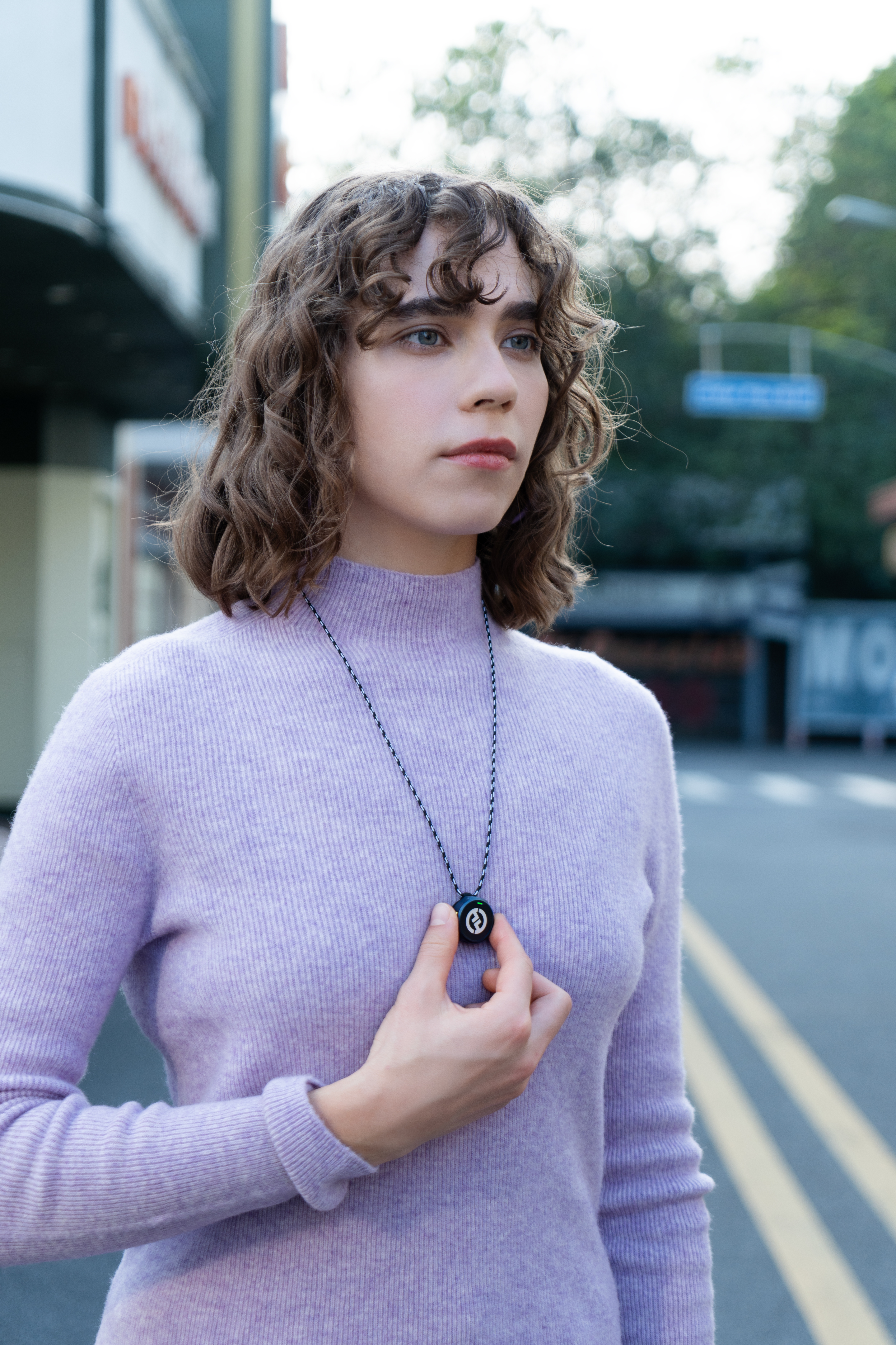 Kobieta w fioletowym swetrze trzyma naszyjnik z czarną zawieszką. Stoi na ulicy.