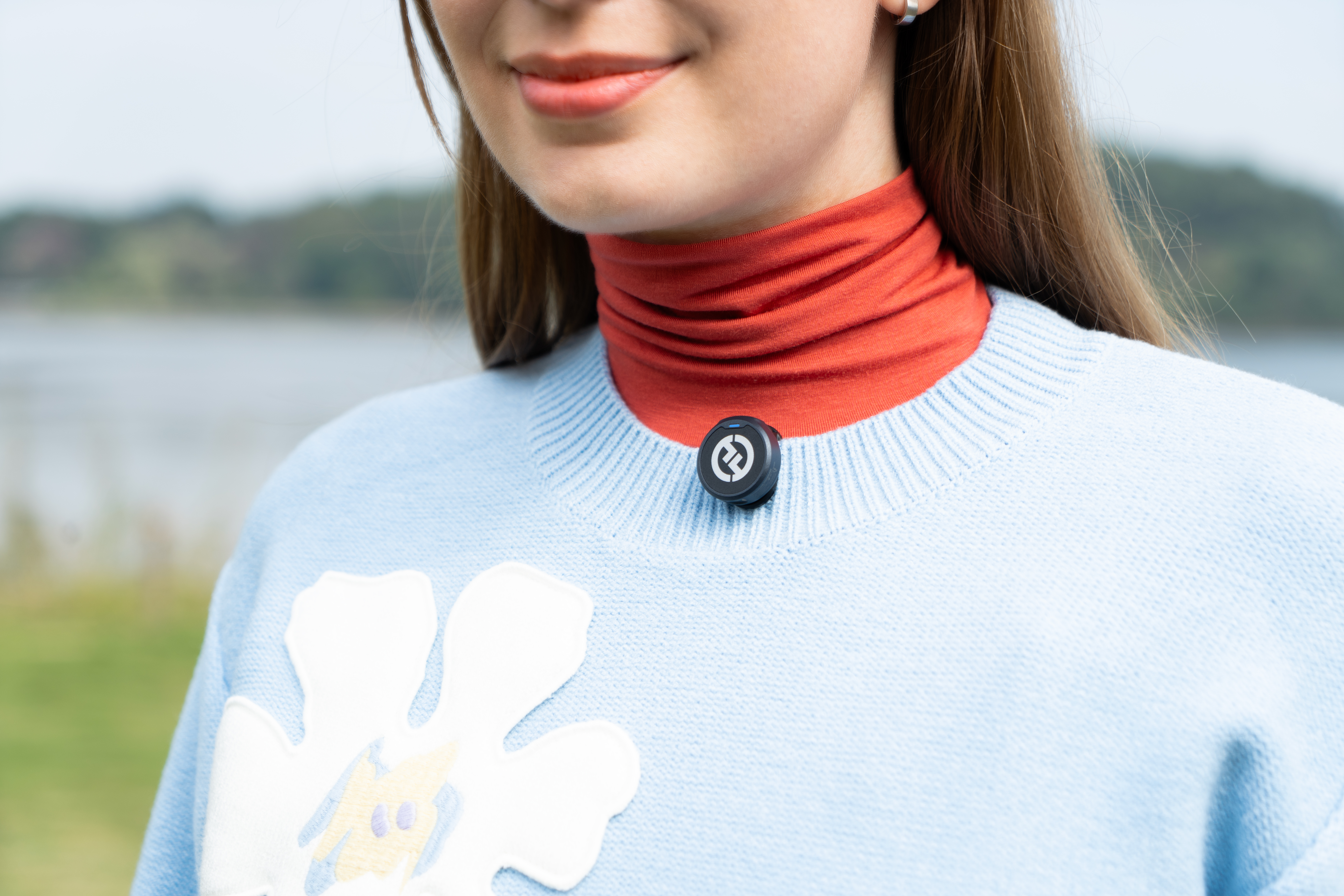 Kobieta w czerwonym golfie pod niebieskim swetrem z białym kwiatem i czarnym urządzeniem.