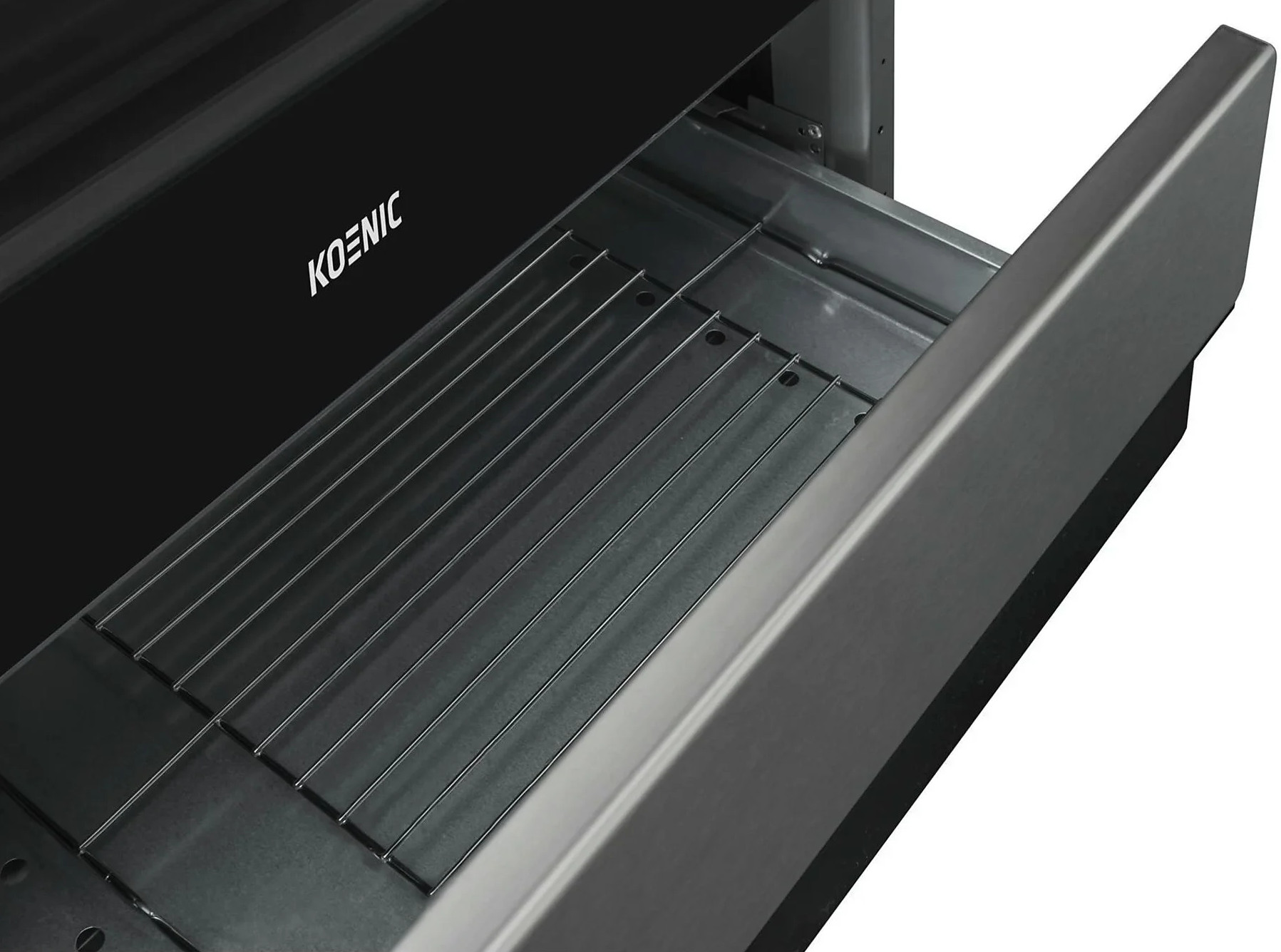 Szuflada piekarnika jest otwarta. Wewnątrz metalowy ruszt znajduje się w metalowej komorze. Czarny front piekarnika ma białą nazwę marki KOENIC.