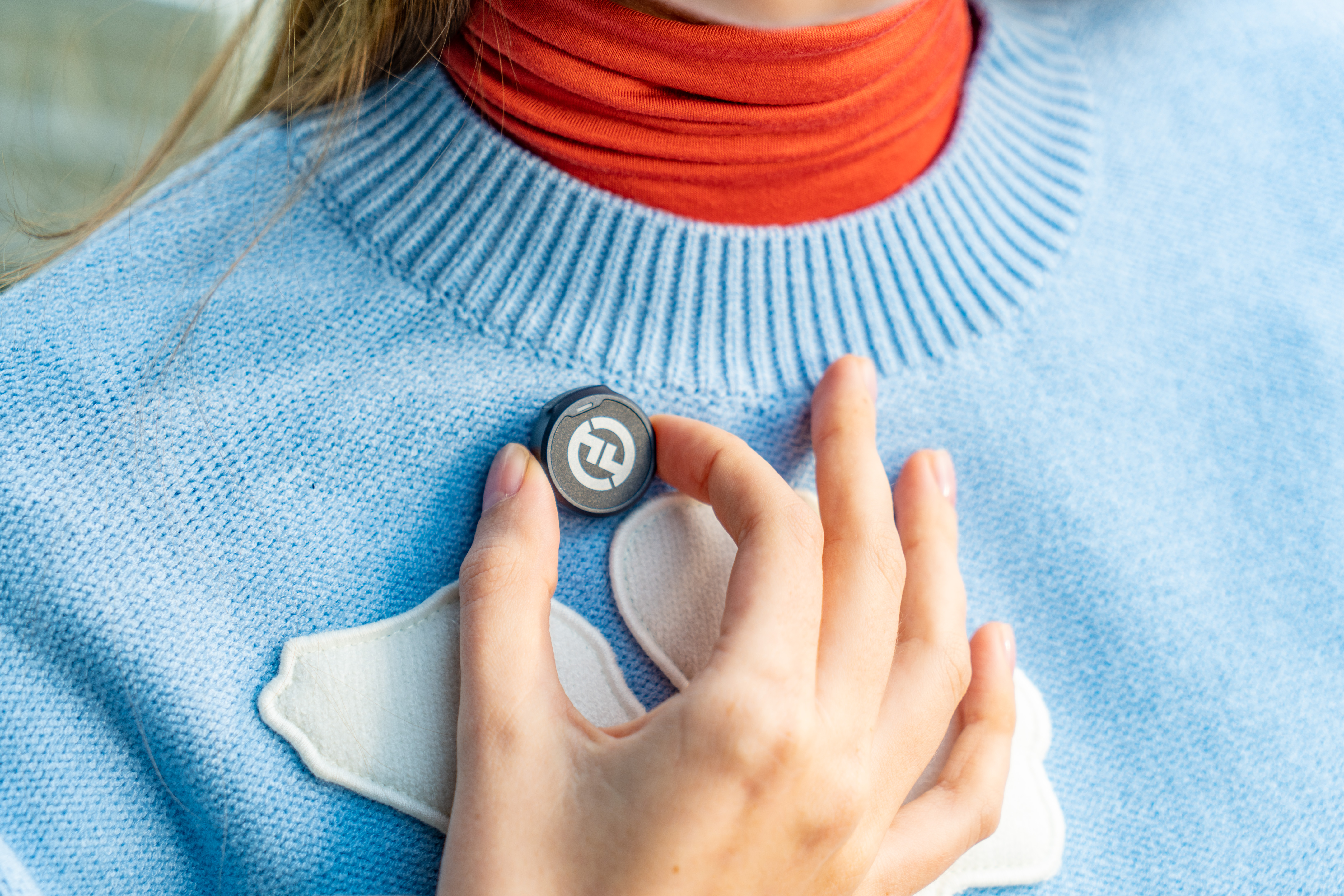 Dłoń trzyma małe, okrągłe, szare urządzenie nad niebieskim swetrem. Widoczny jest pomarańczowy golf.