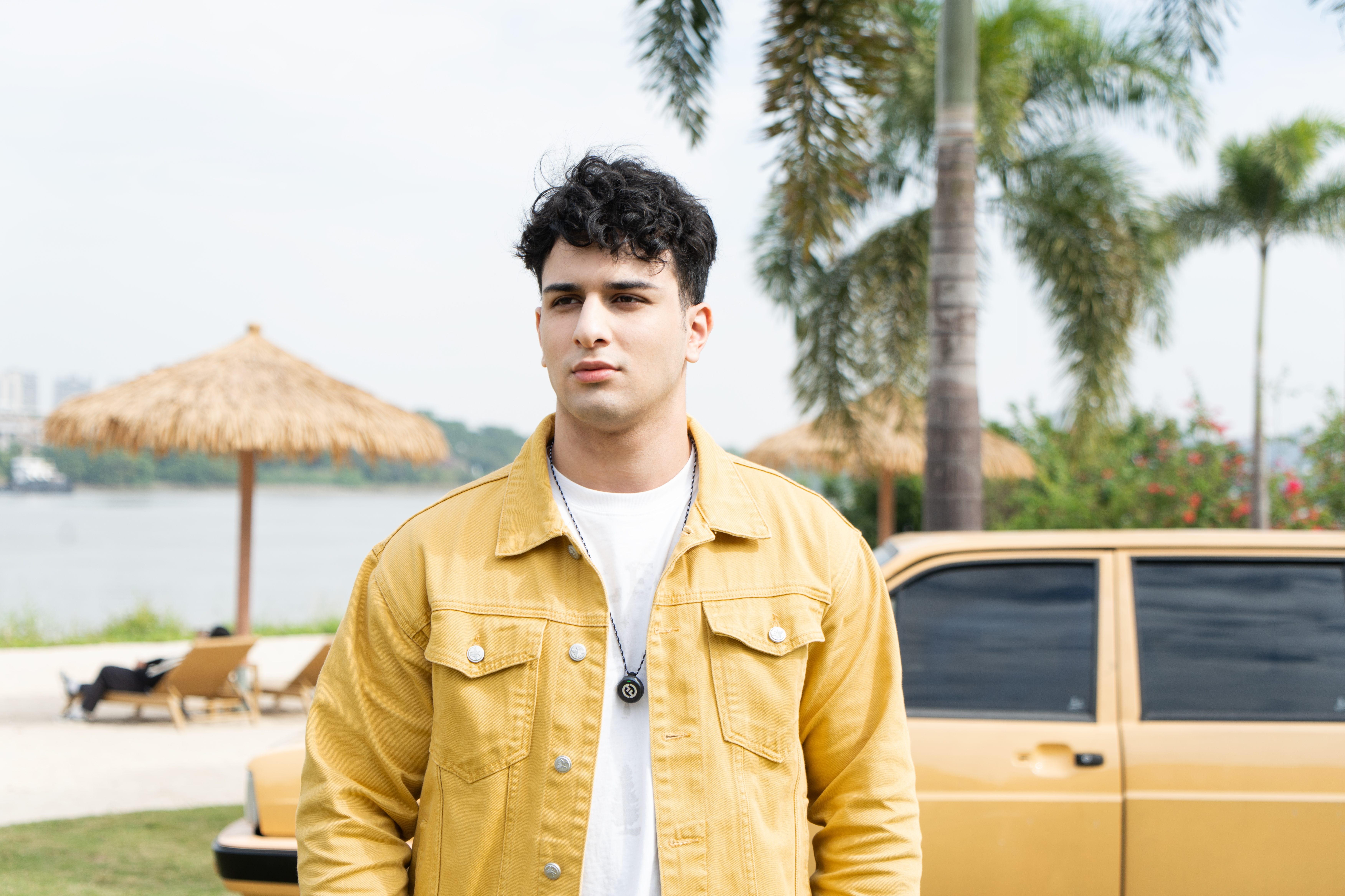 Mężczyzna w żółtej kurtce stoi obok samochodu i palm. W tle parasol ze słomy. Niebo jest czyste.