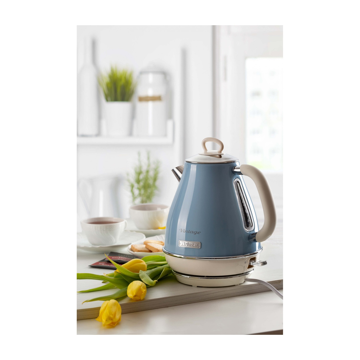 Un bollitore blu vintage su una superficie bianca con tulipani gialli, tazze da tè e biscotti. Scena di cucina luminosa.