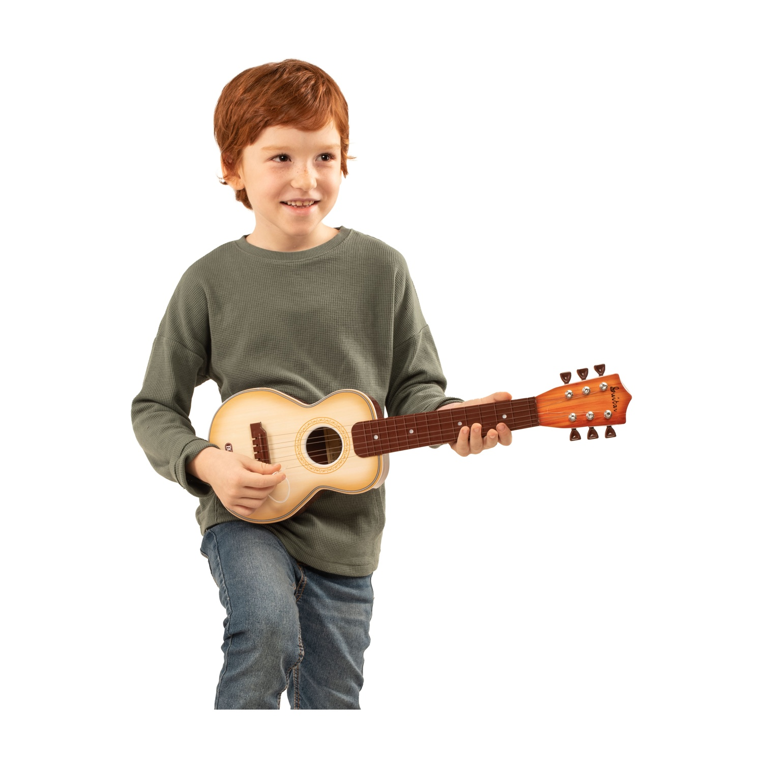 Un ragazzino dai capelli rossi sorride mentre tiene una chitarra giocattolo. Indossa una maglietta verde e jeans, su sfondo bianco.