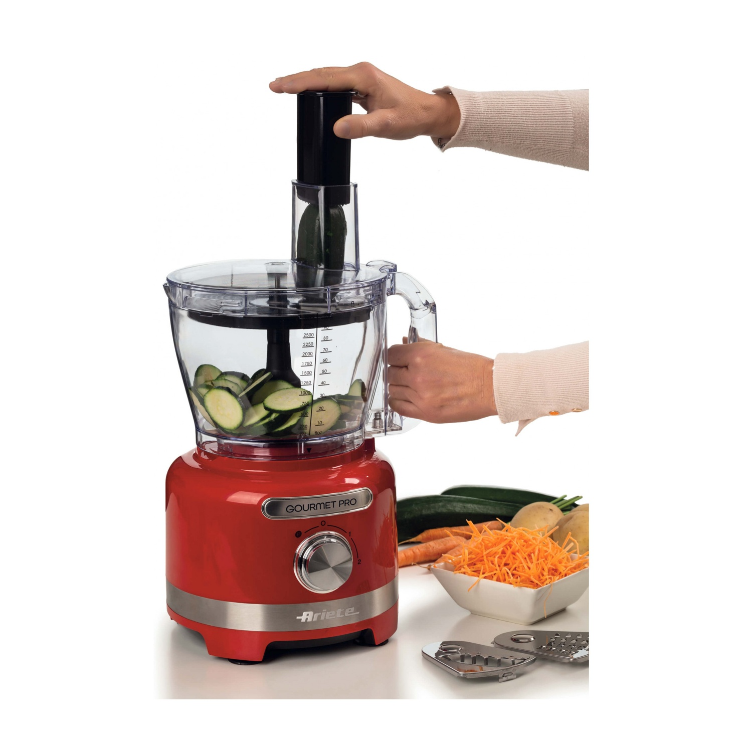 Un robot da cucina rosso con una mano che inserisce una zucchina. Zucchine a fette all'interno del contenitore. Carote grattugiate e altre verdure a lato.