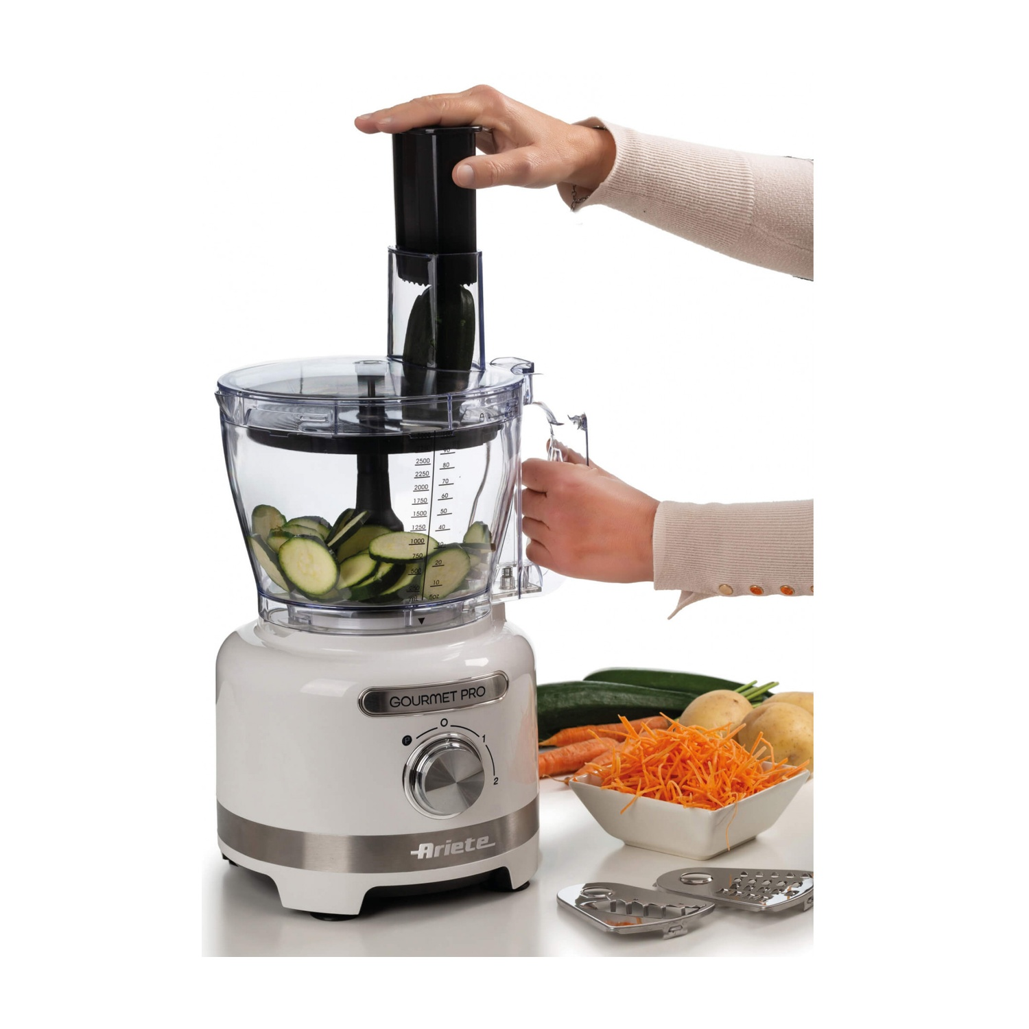 Un robot da cucina con fette di zucchine all'interno. Una mano spinge un pezzo di zucchina nella macchina. Carote e grattugie sul lato.