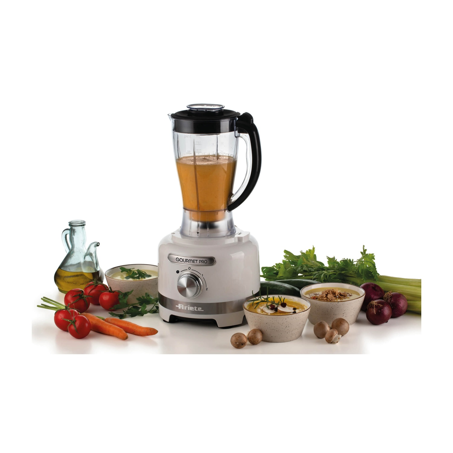Un frullatore con zuppa, circondato da verdure, ciotole di zuppa e olio. Il frullatore è bianco e la zuppa è arancione.