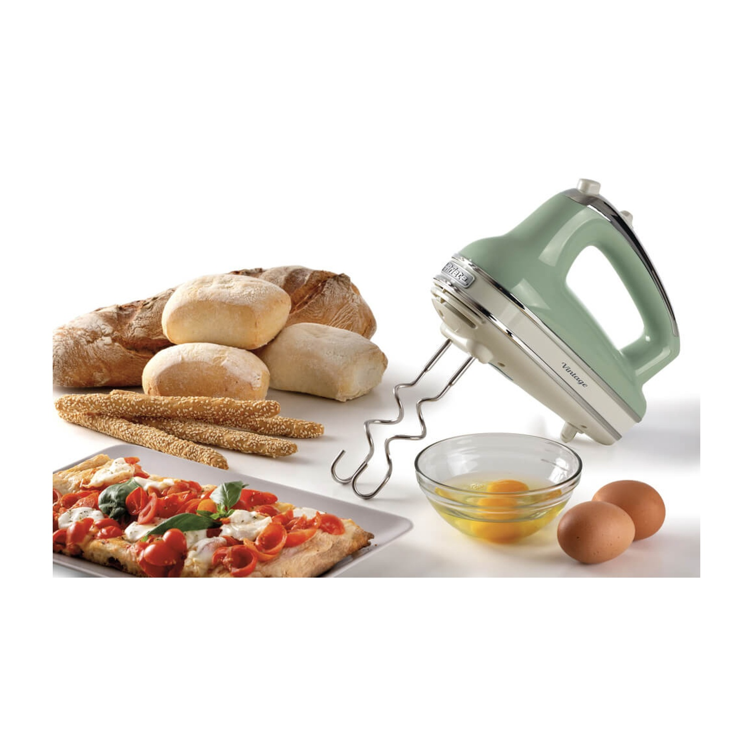 Un mixer elettrico verde menta, pane, pizza, uova e una ciotola con tuorli su una superficie bianca. La scena suggerisce la cucina.