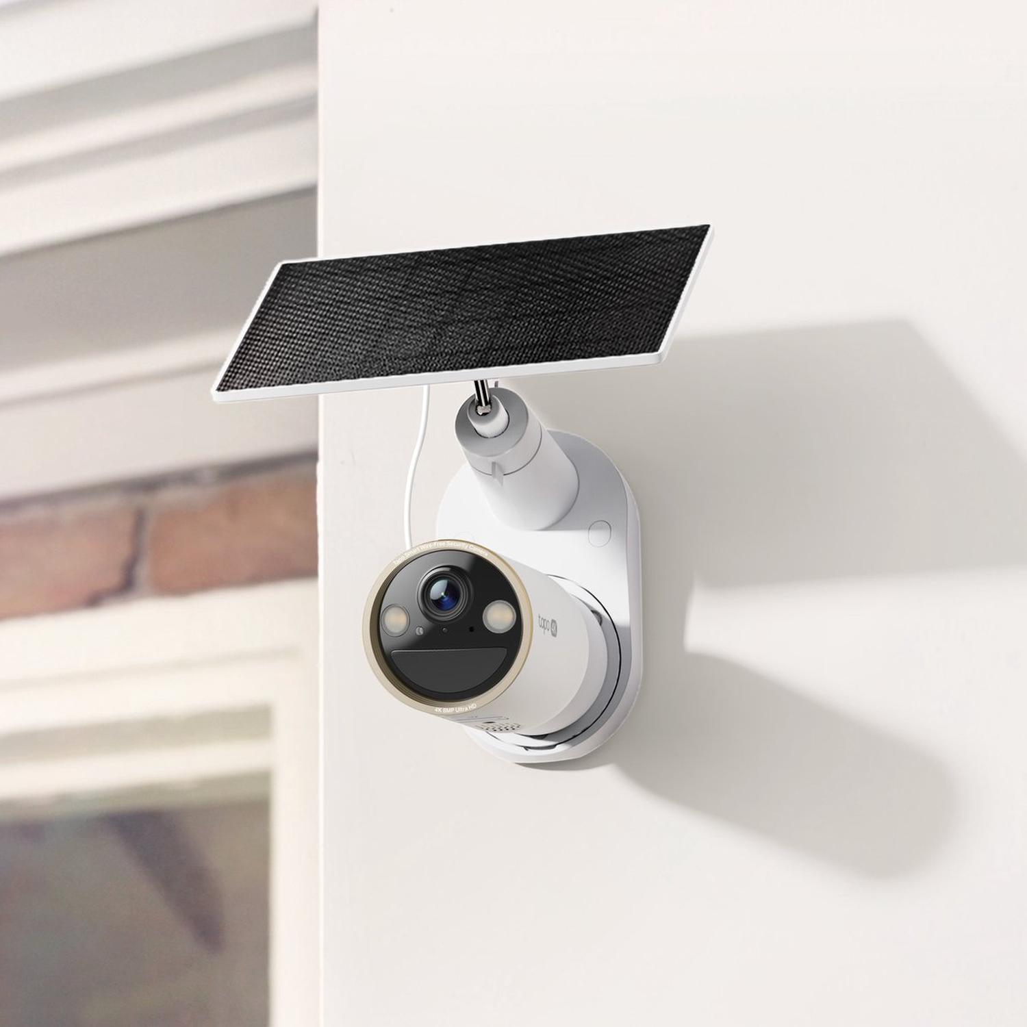 A white security camera with a solar panel is mounted on a white wall. The camera has a black lens and gold ring.