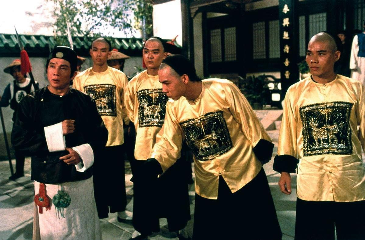 Männer in traditioneller gelber und schwarzer Kleidung. Einige sind kahl. Szene hat eine traditionelle chinesische Architektur im Hintergrund.
