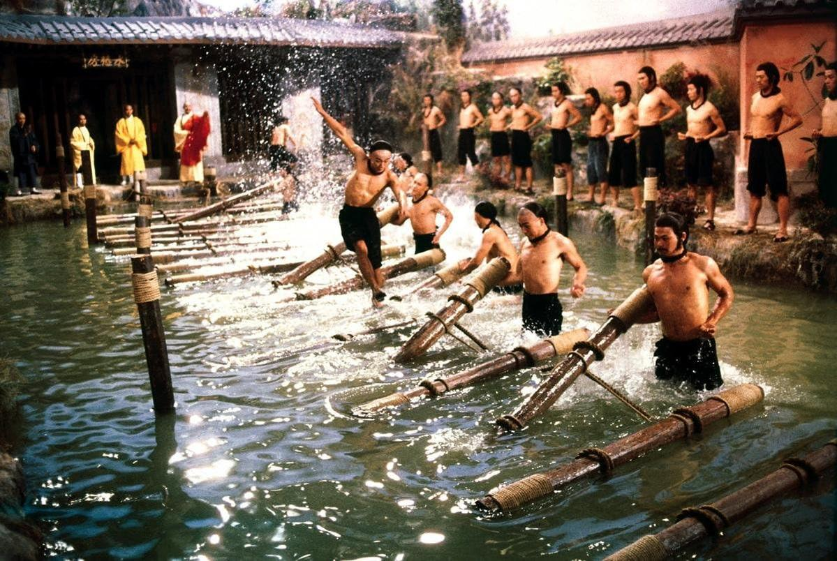 Männer trainieren im Wasser. Einige sind ohne Hemd, andere auf einer Holzbrücke. Im Hintergrund ein Gebäude und Zuschauer.