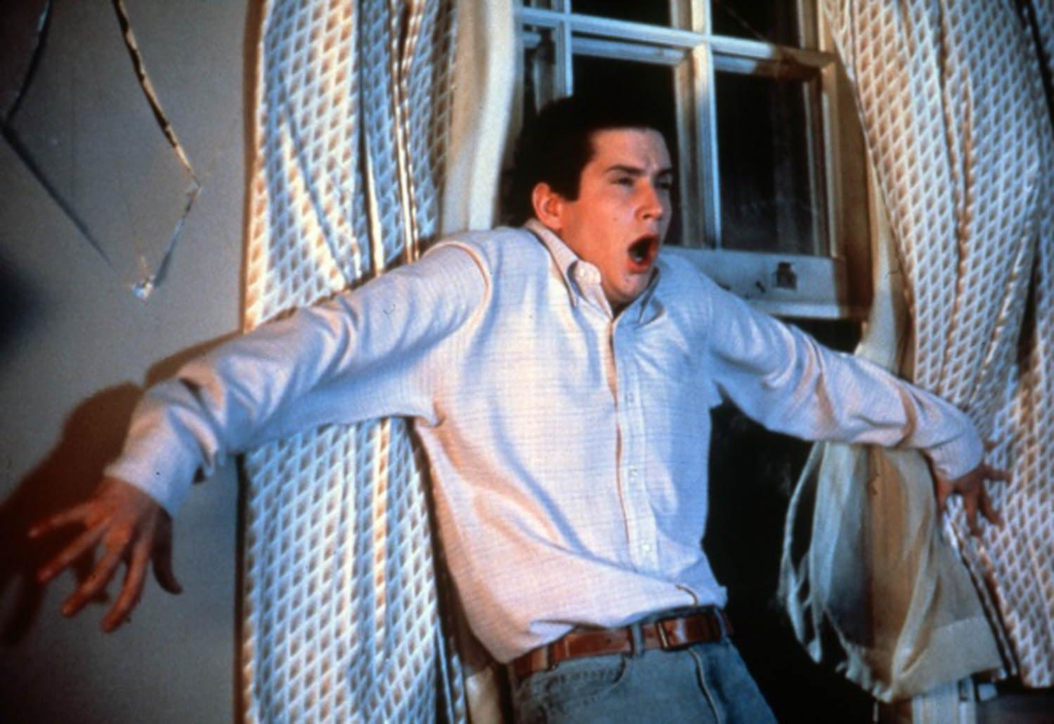 Mann mit offenem Mund, Arme ausgestreckt, in hellblauem Hemd und Jeans. Fenster und Vorhänge im Hintergrund.