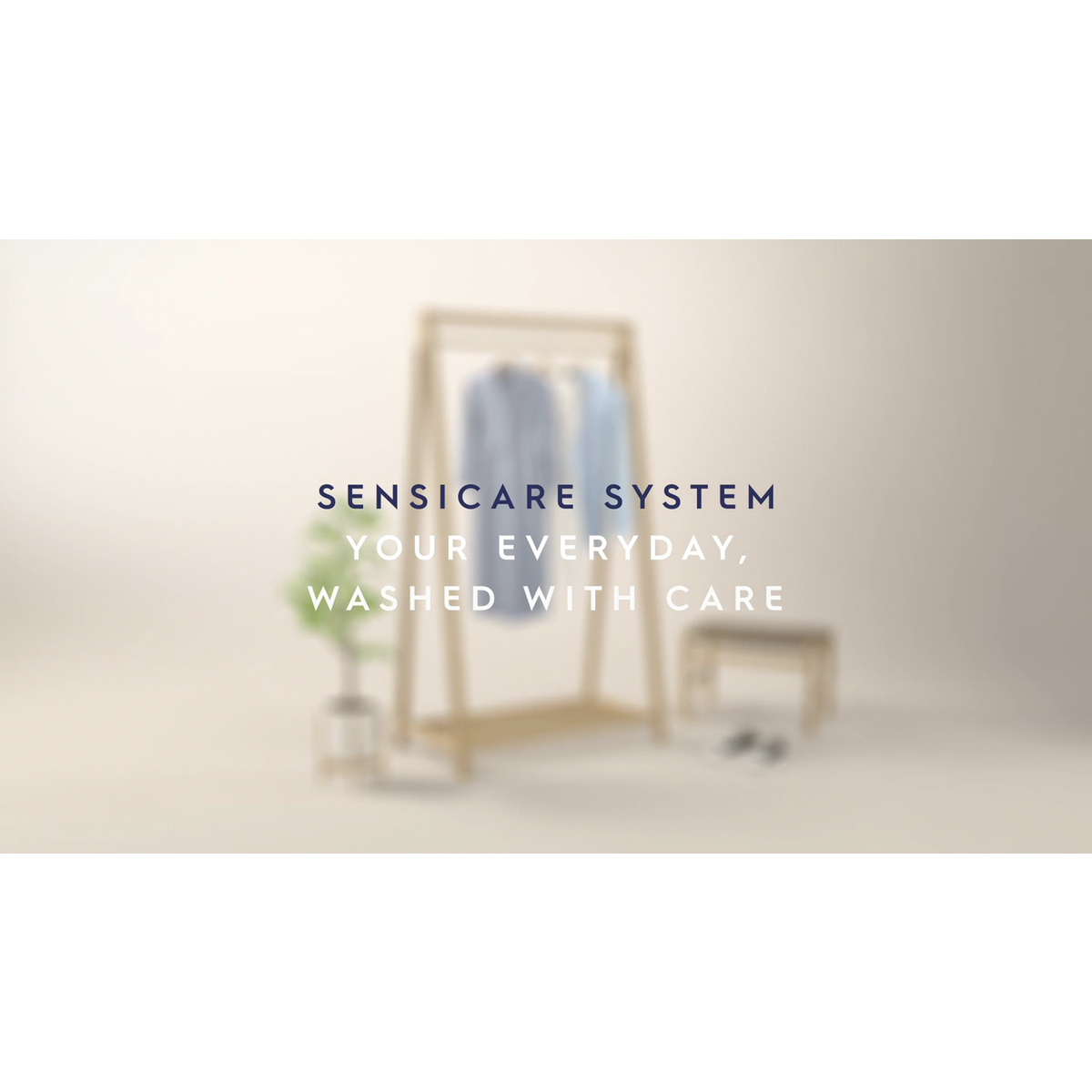 Elmosódott kép szöveggel. Egy fából készült ruhakampó ingekkel. Egy kis növény és egy zsámoly a közelben. Szöveg: SENSICARE SYSTEM, washed with care.