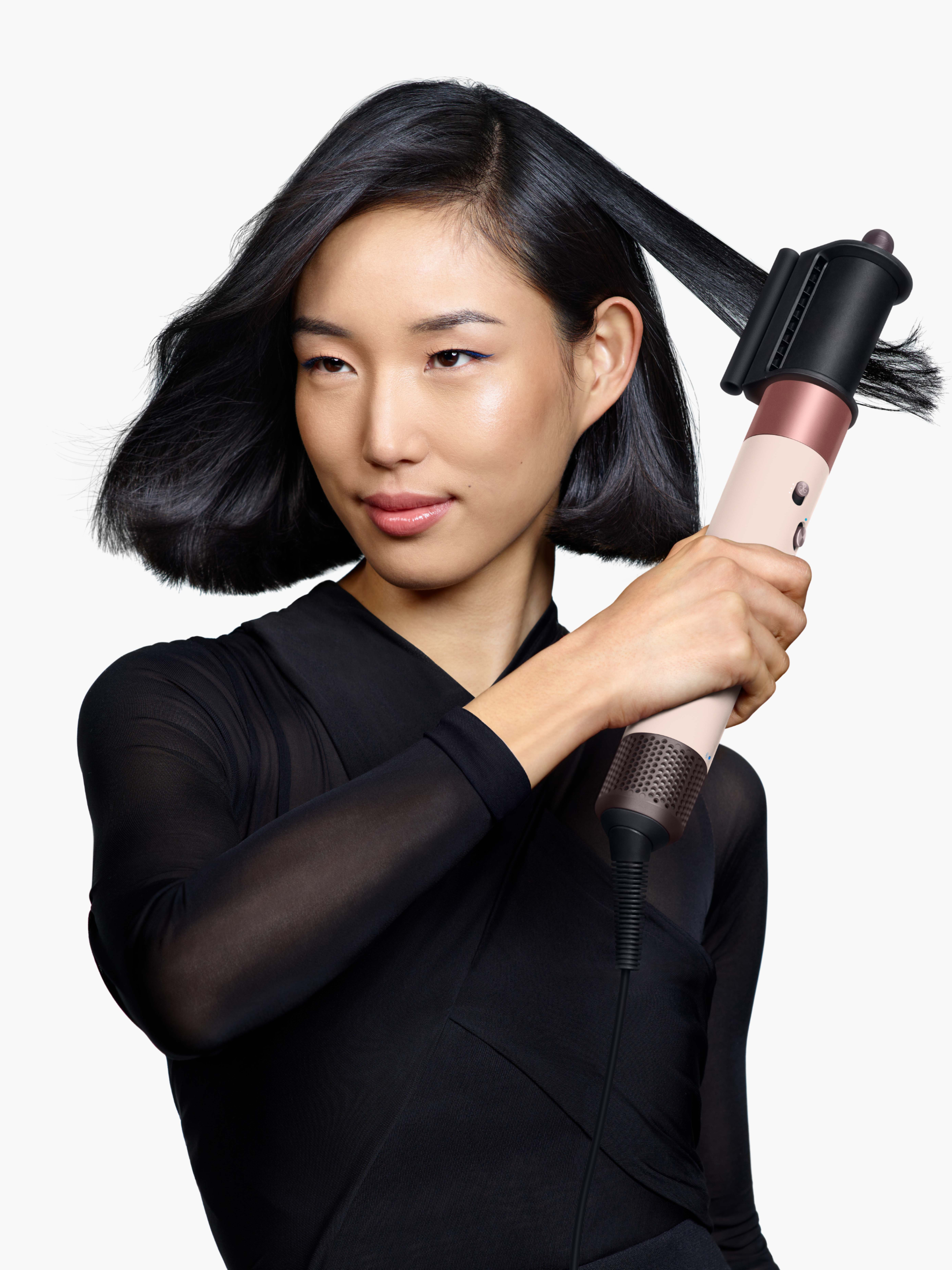 Mujer peinando su cabello negro con un secador. Viste ropa negra. Fondo blanco.