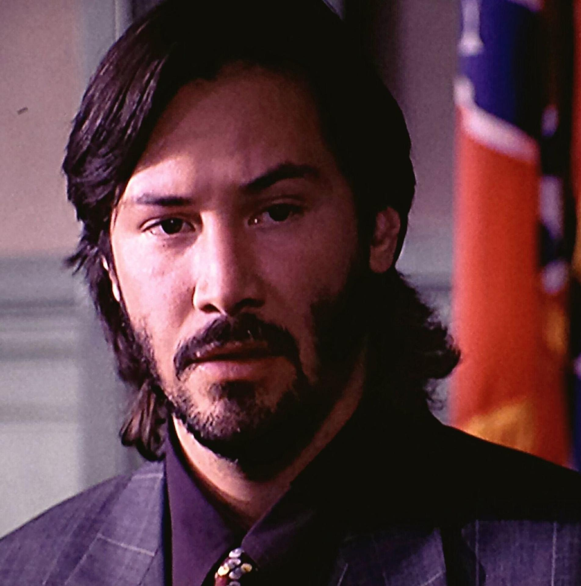 Nahaufnahme von Keanu Reeves mit Bart und langem Haar, trägt einen Anzug. Im Hintergrund ist die amerikanische Flagge zu sehen.