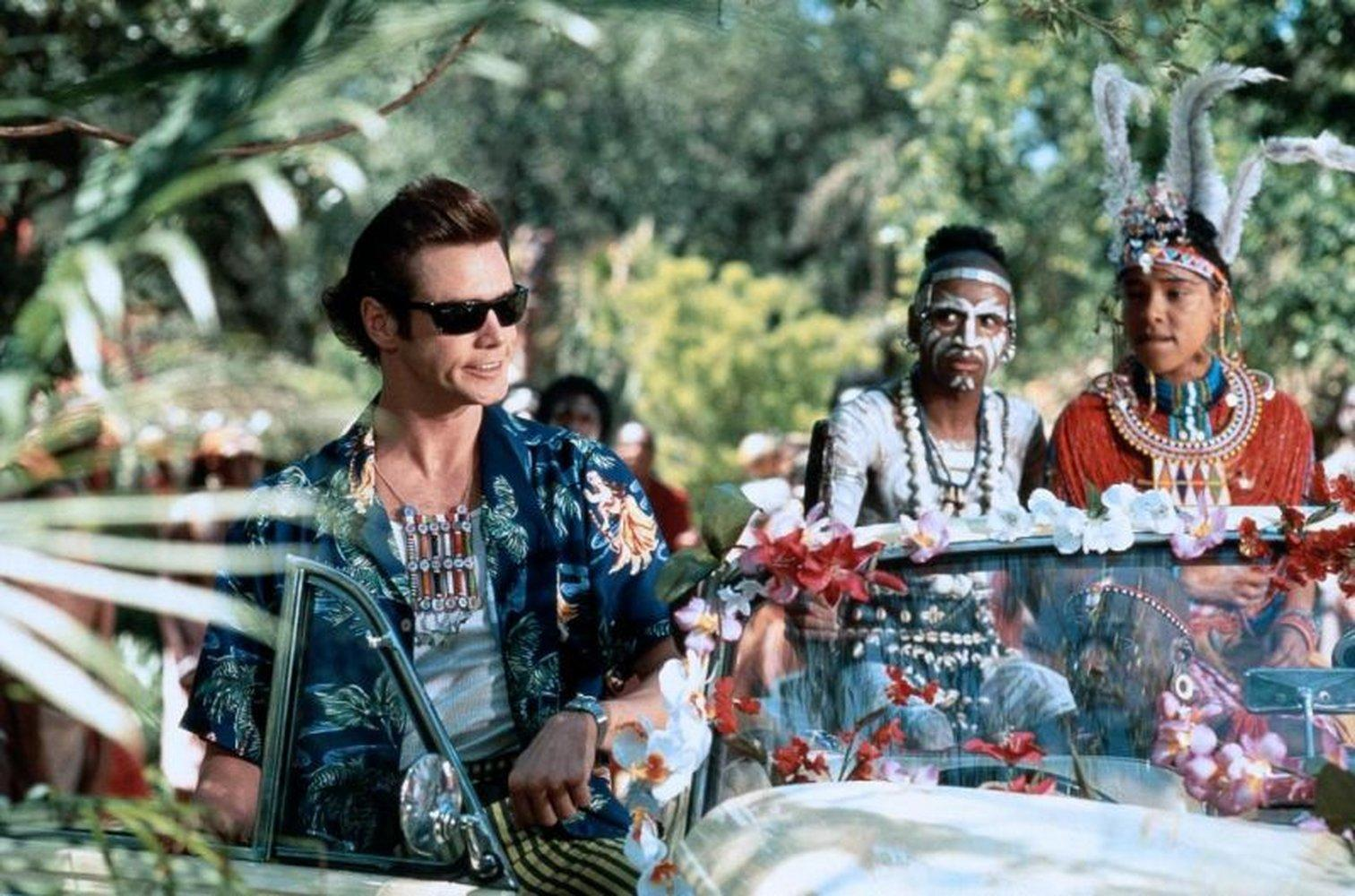 Jim Carrey mit Sonnenbrille in einem Auto mit Menschen in Stammeskleidung. Blumen schmücken das Auto. Grünes Laub im Hintergrund.