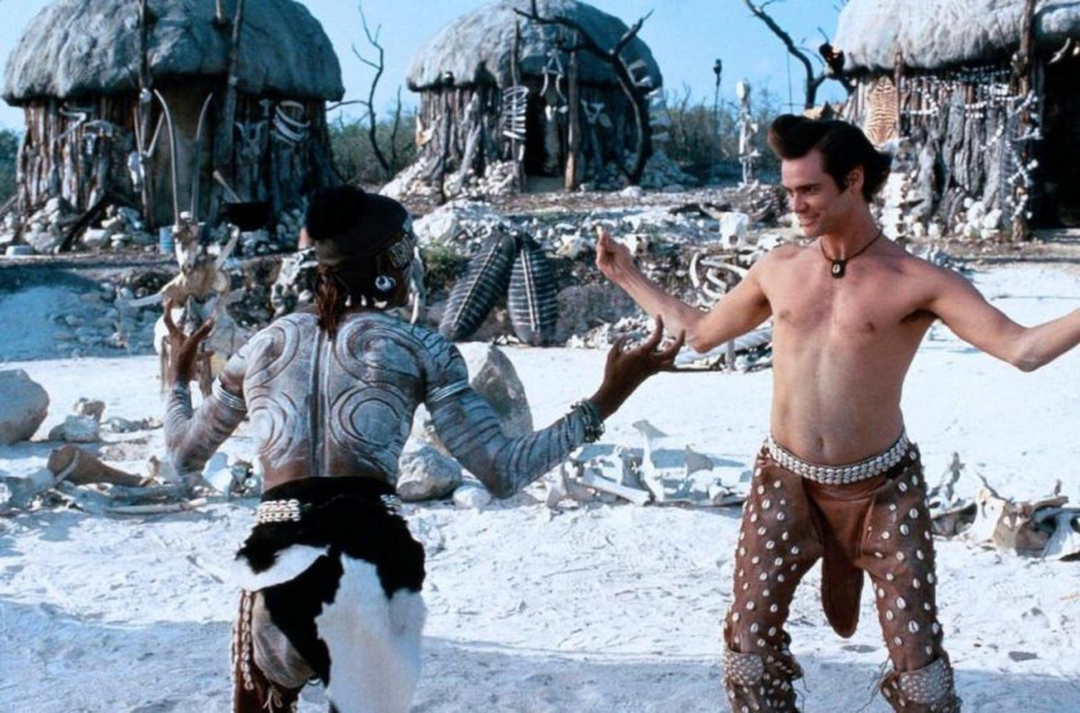 Jim Carrey und eine andere Person in Stammeskleidung. Sie stehen auf weißem Sand, im Hintergrund Hütten.