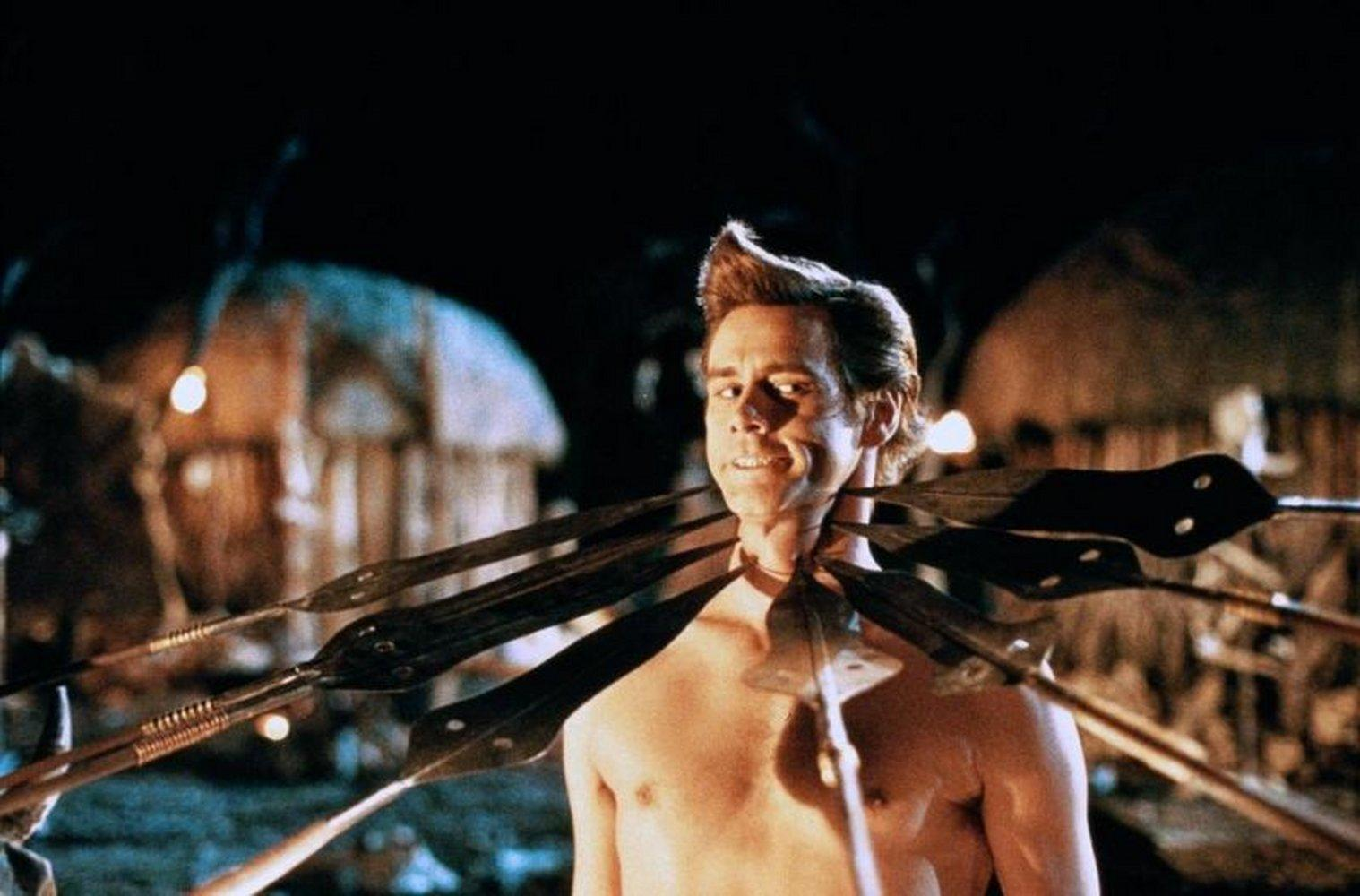 Jim Carrey als Ace Ventura mit Speeren um den Hals, lächelnd. Dunkler Hintergrund mit Hütten und Lichtern.