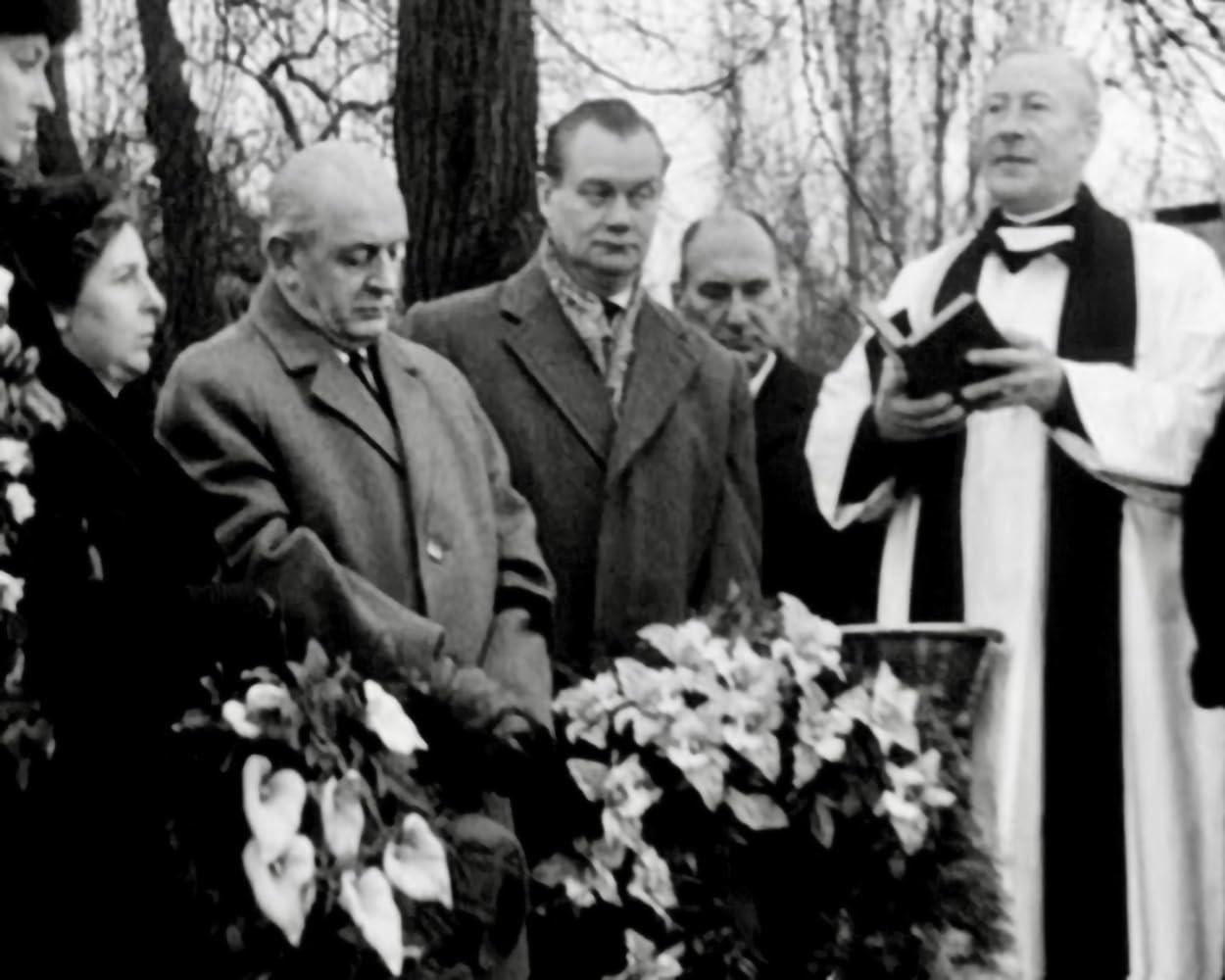 Eine Gruppe von Menschen bei einer Beerdigung. Ein Priester liest aus einem Buch vor. Blumen sind im Vordergrund.