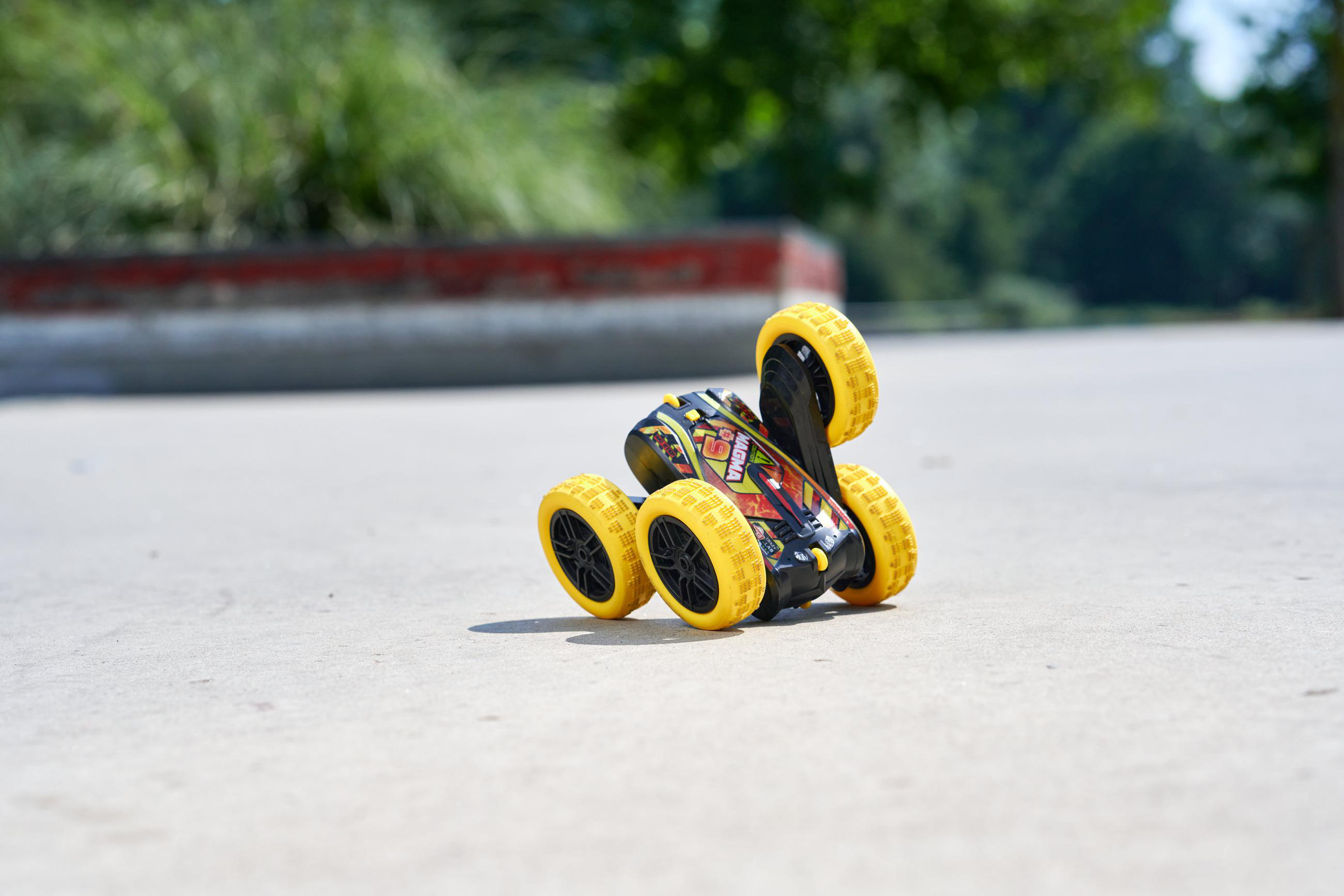 Ein Spielzeugauto mit gelben Rädern steht auf einer Betonoberfläche. Im Hintergrund sind grüne Bäume.