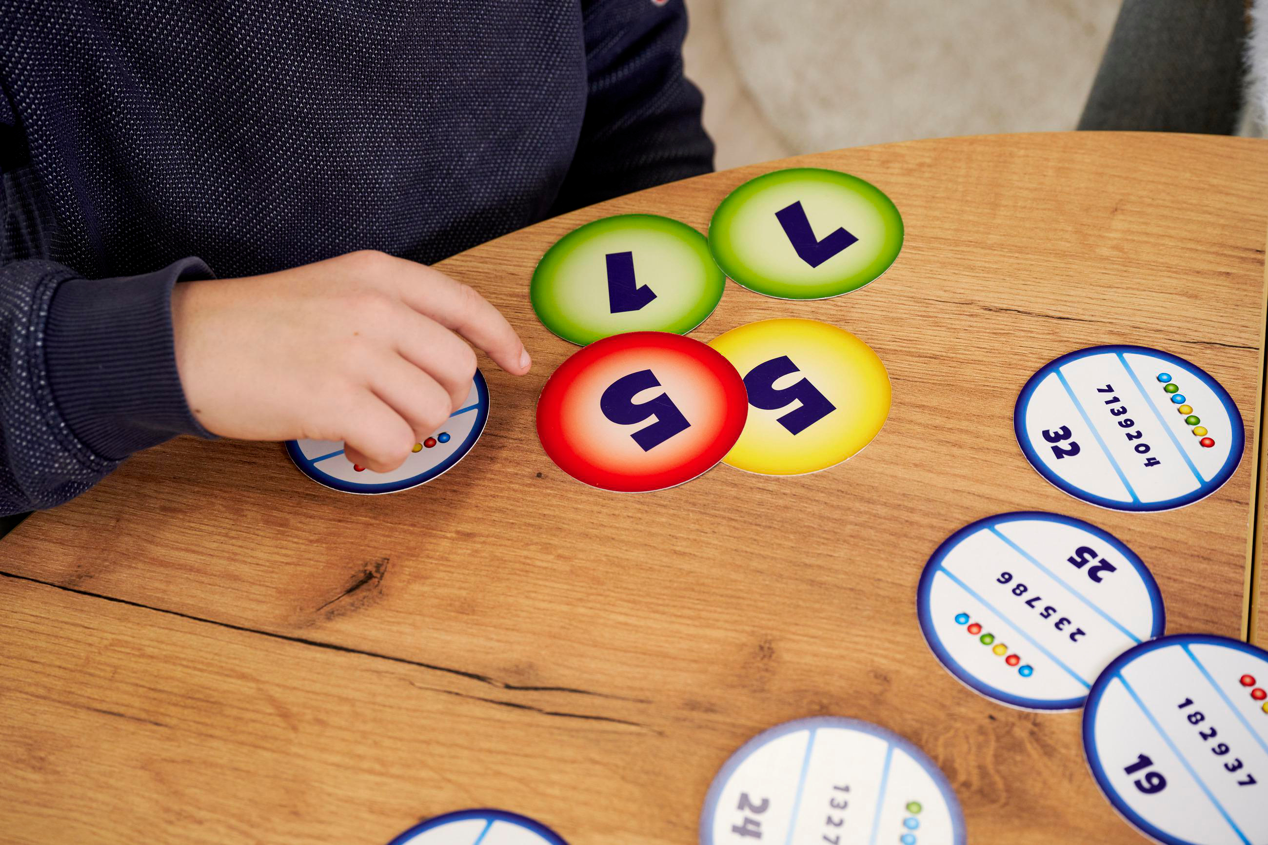 Die Hand einer Person berührt ein Spielstück. Mehrere bunte Spielsteine mit Zahlen und ein Holztisch sind sichtbar.