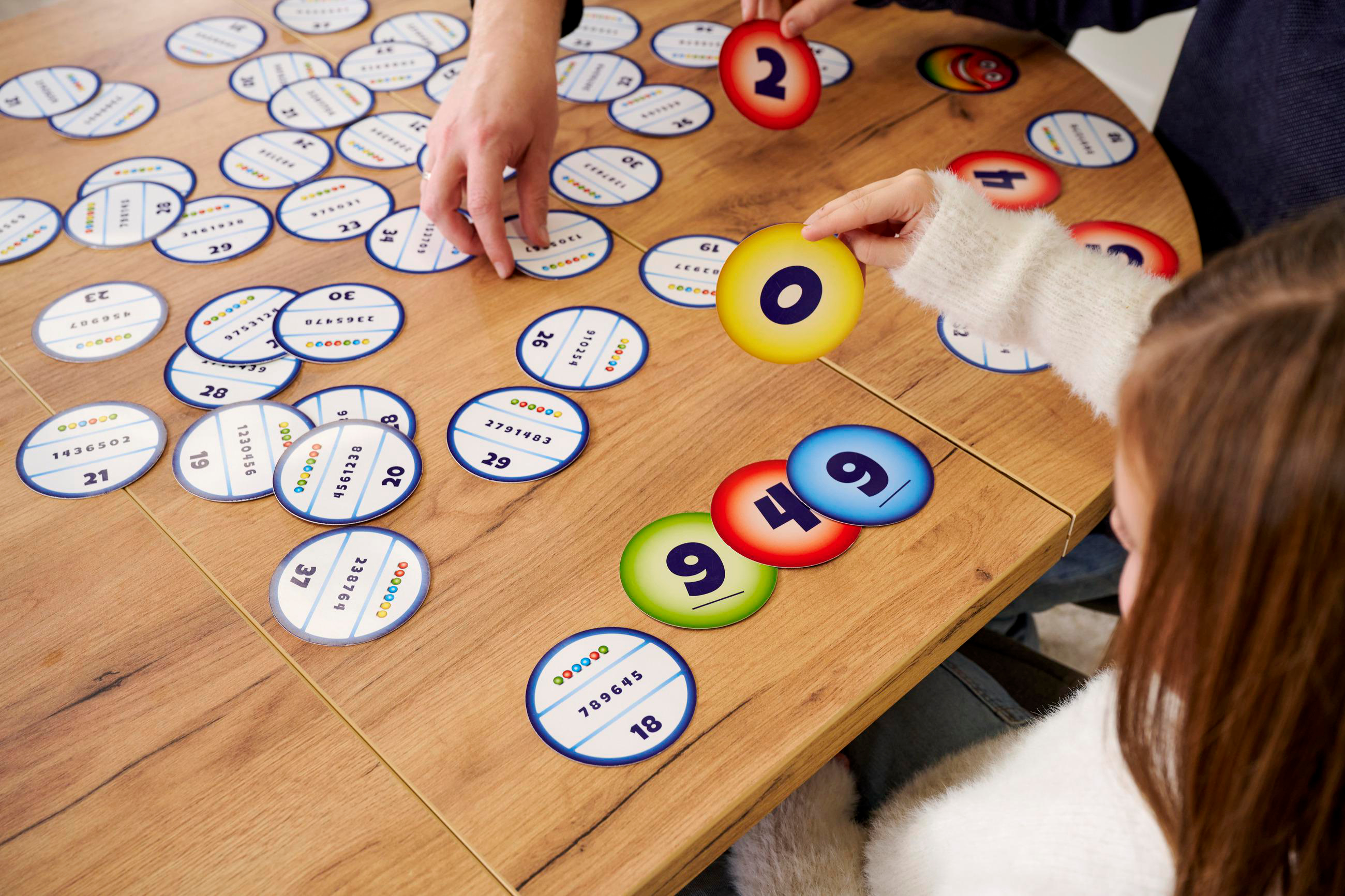 Leute spielen ein Zahlenspiel auf einem Holztisch. Hände halten farbige Zahlenkarten und Spielsteine.