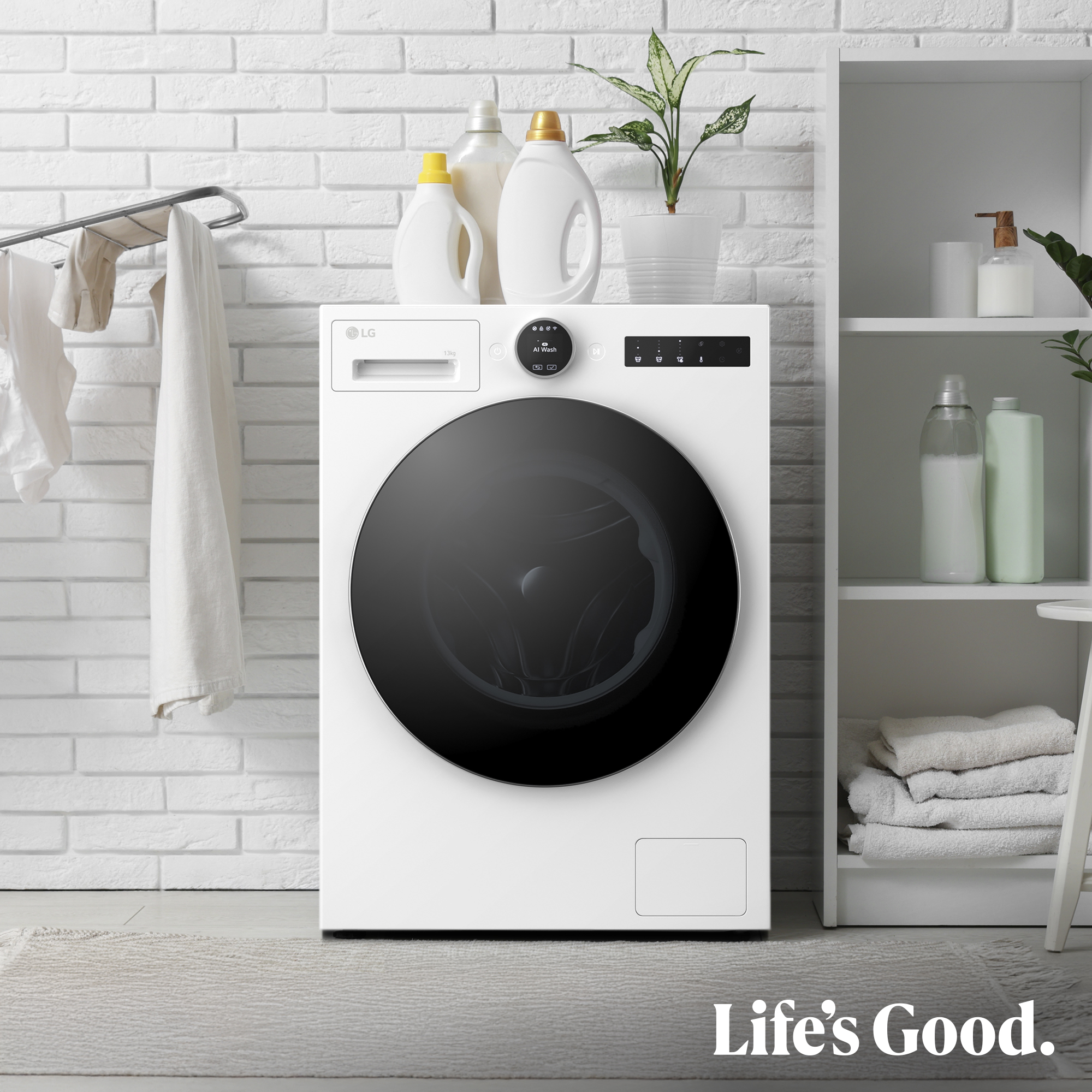 Una lavadora LG blanca en un lavadero. Productos de lavandería, una planta y toallas completan la escena. El lema: Life's Good.