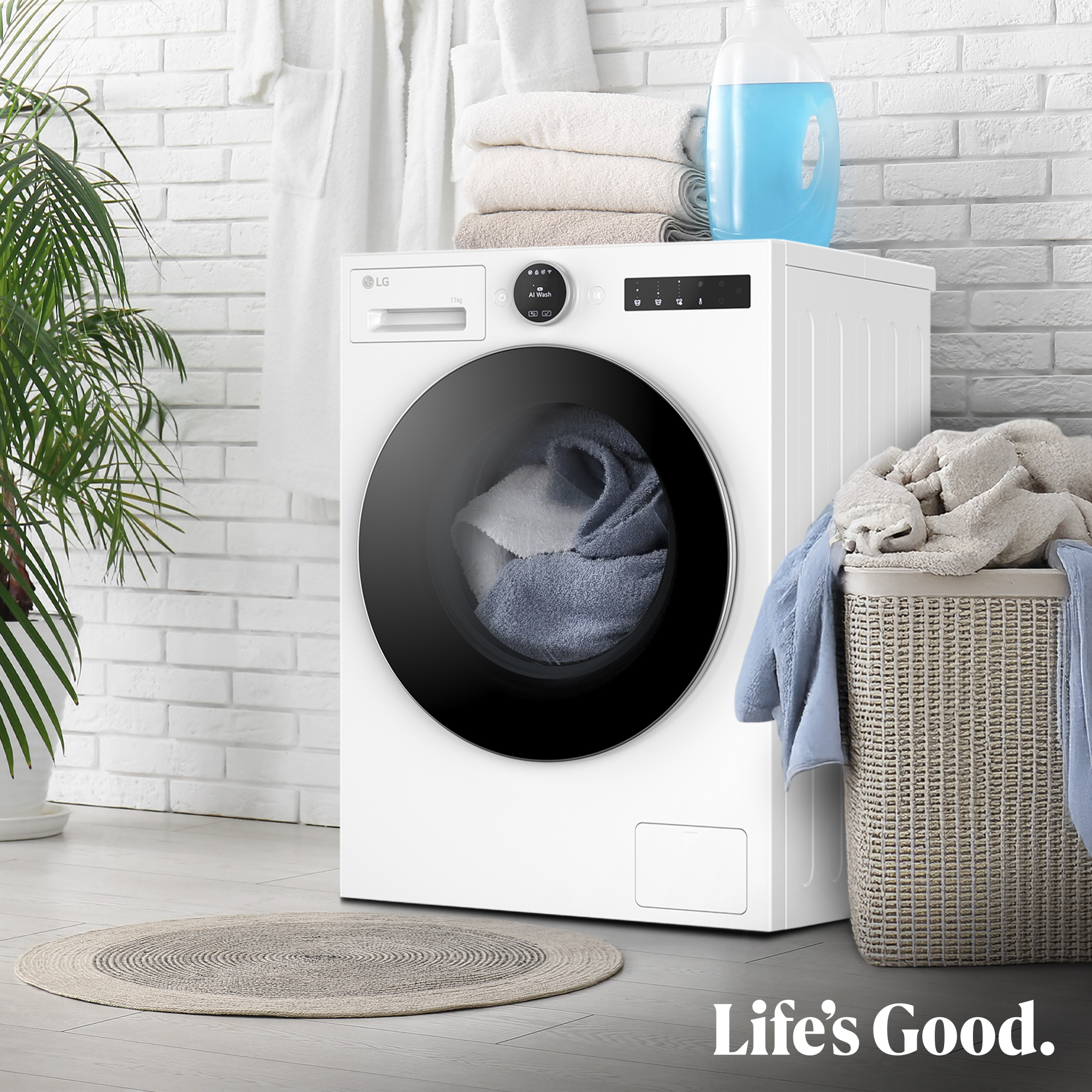 Una lavadora LG blanca con toallas, cesta de ropa y una planta está en un baño. Life's Good está escrito abajo.