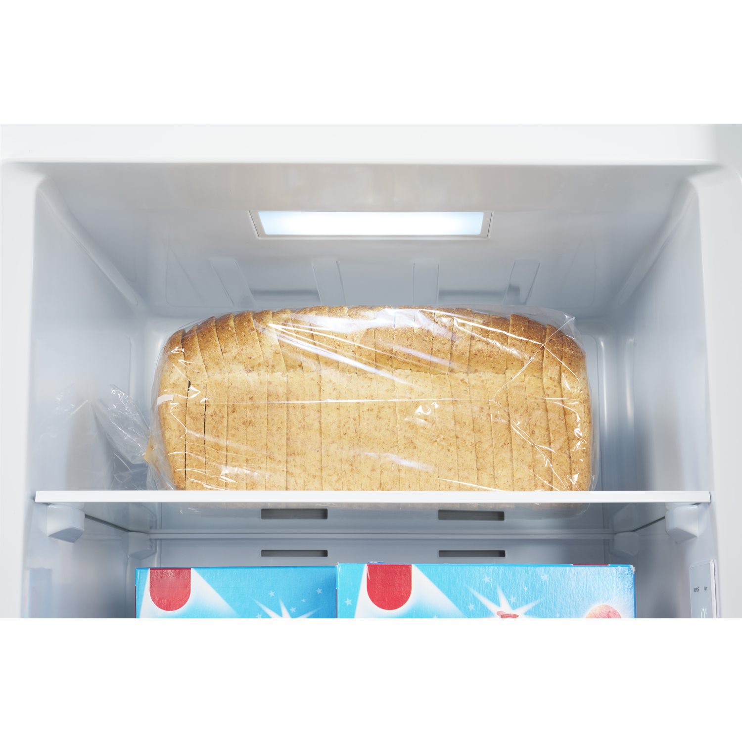 A loaf of sliced bread in a clear plastic bag sits on a shelf inside a refrigerator. Below, are two boxes.