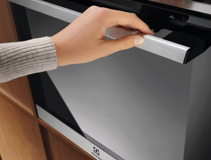 Ręka otwiera drzwi piekarnika. Piekarnik jest czarny ze srebrnym uchwytem. Widoczne jest logo Electrolux.