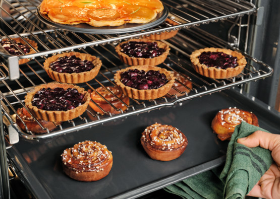 Piekarnik wypełniony wypiekami: tartami, bułeczkami cynamonowymi i ciastem. Ręka trzyma zieloną szmatkę.