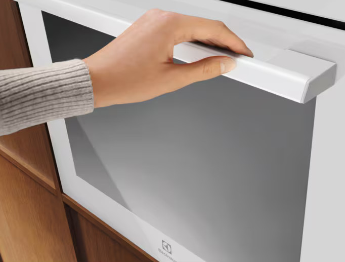 Ręka otwiera białe drzwi piekarnika. Piekarnik ma szklany front i jest umieszczony w drewnianej szafce. Osoba ma na sobie szary sweter.