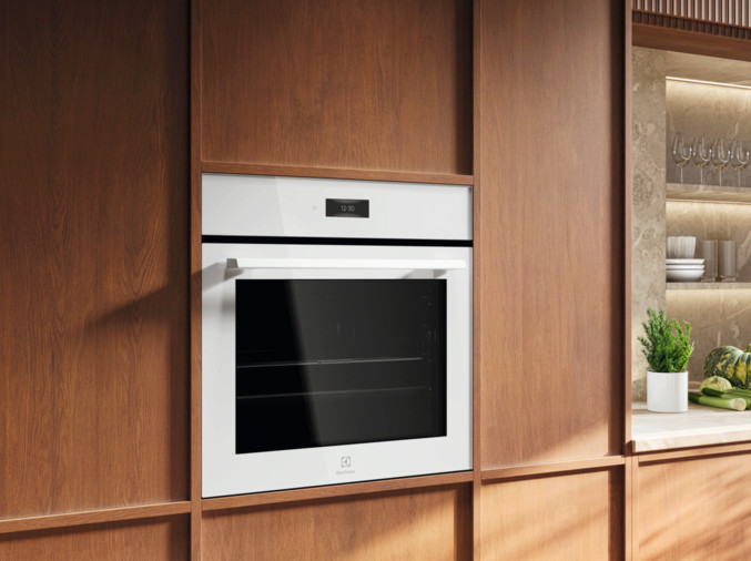 Biały piekarnik do zabudowy jest obramowany drewnianymi panelami. Wyświetlacz pokazuje 12:30. W tle blat z roślinami i akcesoriami kuchennymi.