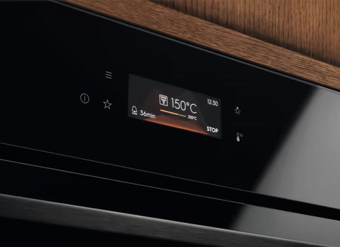 Czarny panel sterowania piekarnika pokazujący 150°C, 36 minut i 12:30. Powierzchnia drewniana powyżej. Ikony i przyciski widoczne.