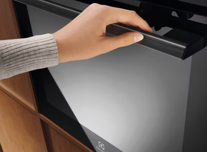 Ręka otwiera drzwi piekarnika. Piekarnik jest czarny, a uchwyt ciemnoszary. Ręka jest blada, w szarym swetrze.