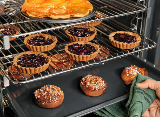 Piekarnik z wypiekami: tarty z jagodami, cynamonki i ciasto. Ręka trzyma zieloną szmatkę.