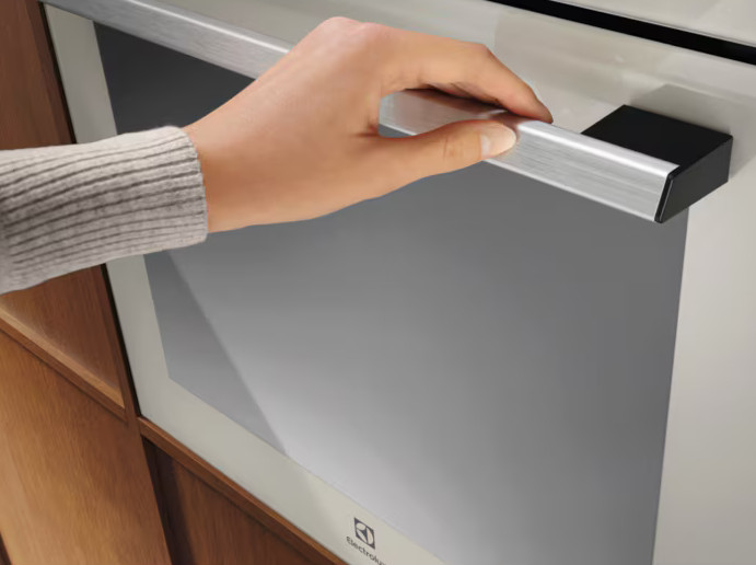 Ręka trzyma metalowy uchwyt na białych drzwiach piekarnika. Piekarnik ma szary szklany front. Widoczne są drewniane szafki.