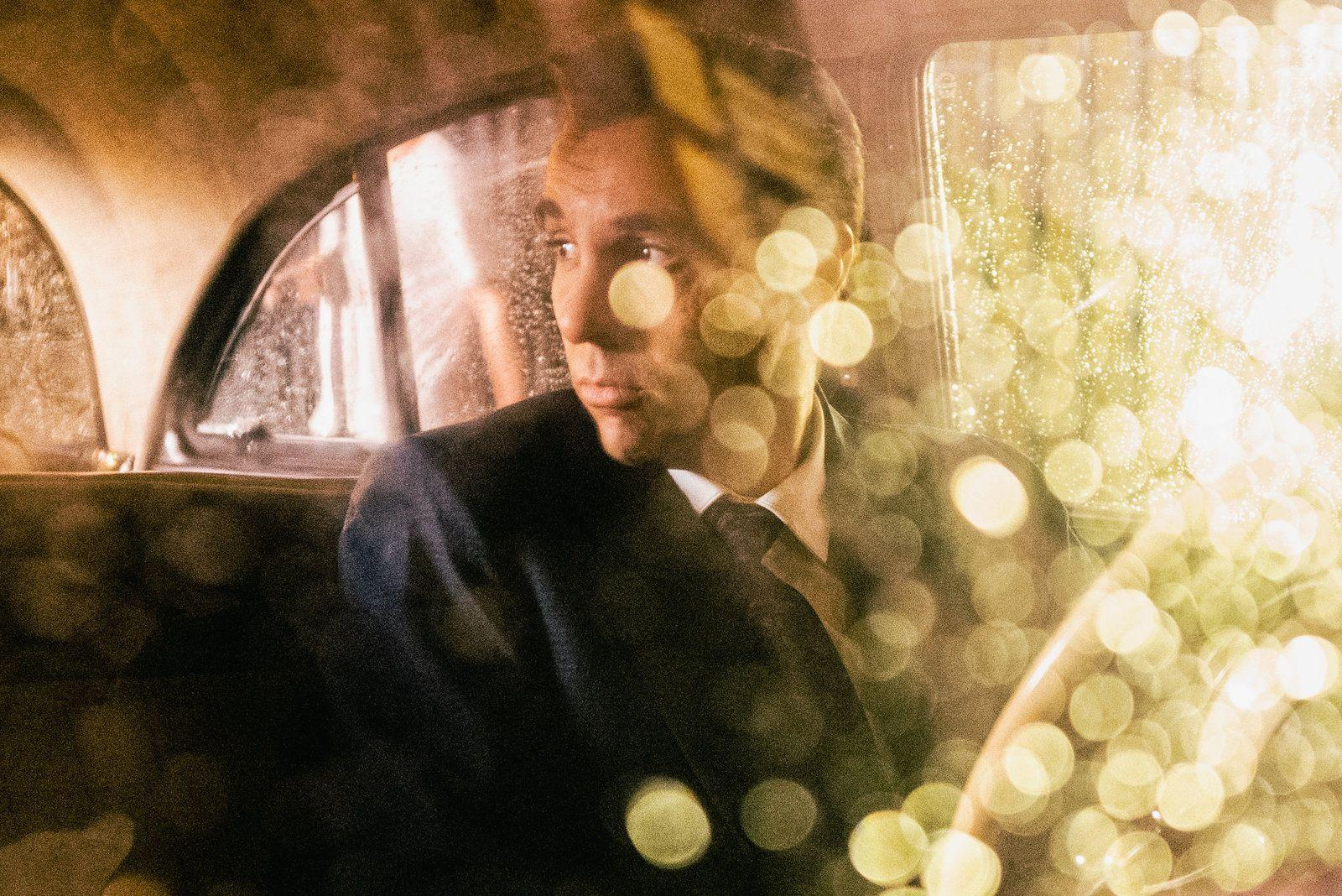 Ein Mann im Anzug schaut aus einem Autofenster. Regen zieht über das Glas. Bokeh-Lichter überlagern die Szene und erzeugen ein sanftes Leuchten.
