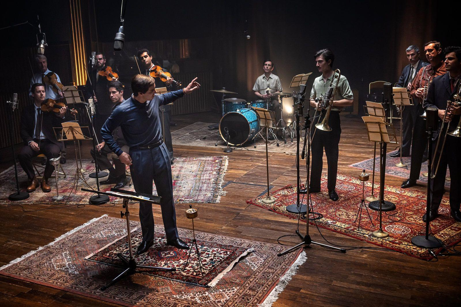 Eine Band spielt in einem Studio. Musiker spielen Geige, Schlagzeug und Blechbläser. Ein Dirigent dirigiert von der Mitte aus, Mikrofone herum.