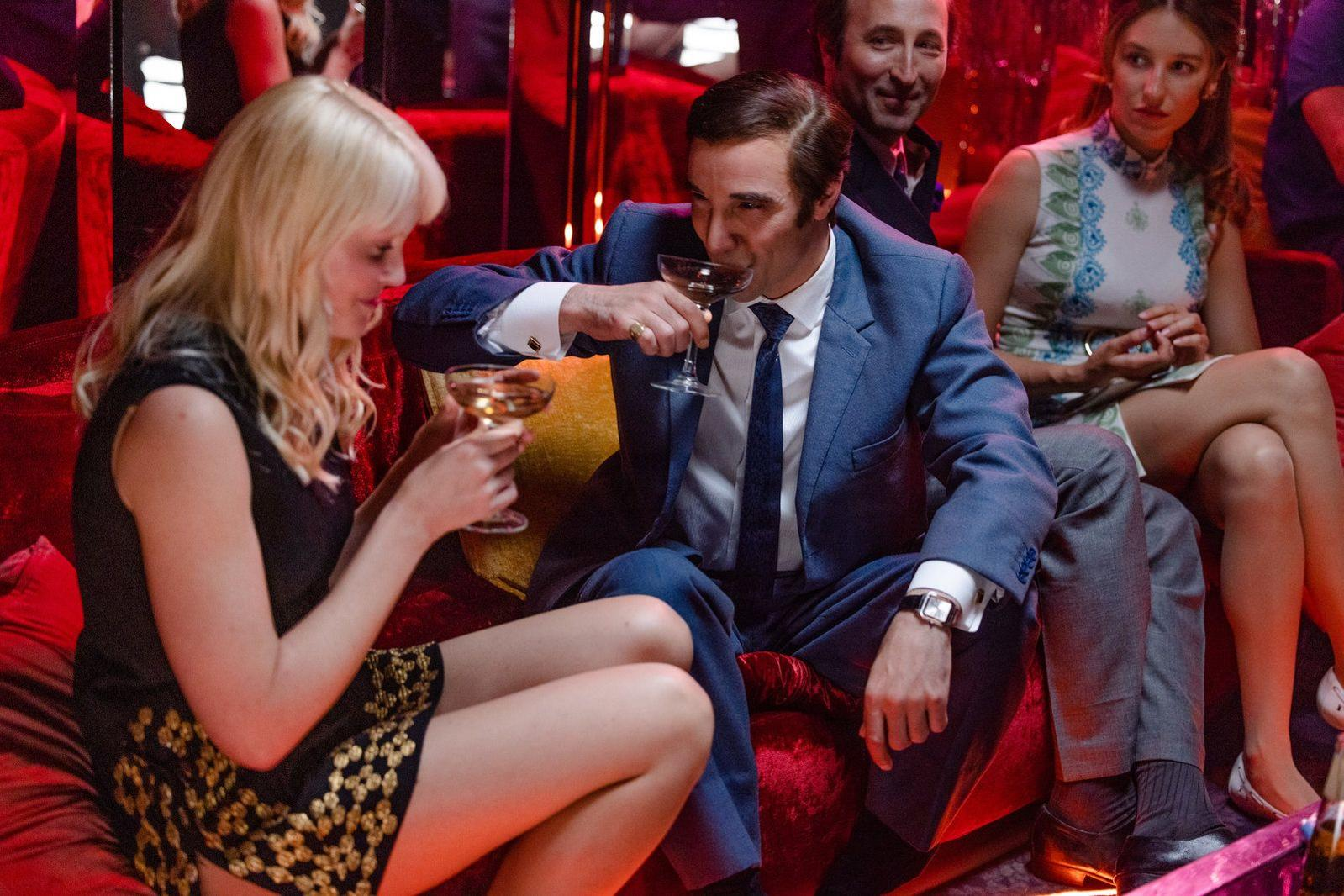 Ein Mann im blauen Anzug trinkt aus einem Glas. Eine blonde Frau und zwei andere sind in der Nähe. Die Szene spielt in einer schwach beleuchteten Lounge.