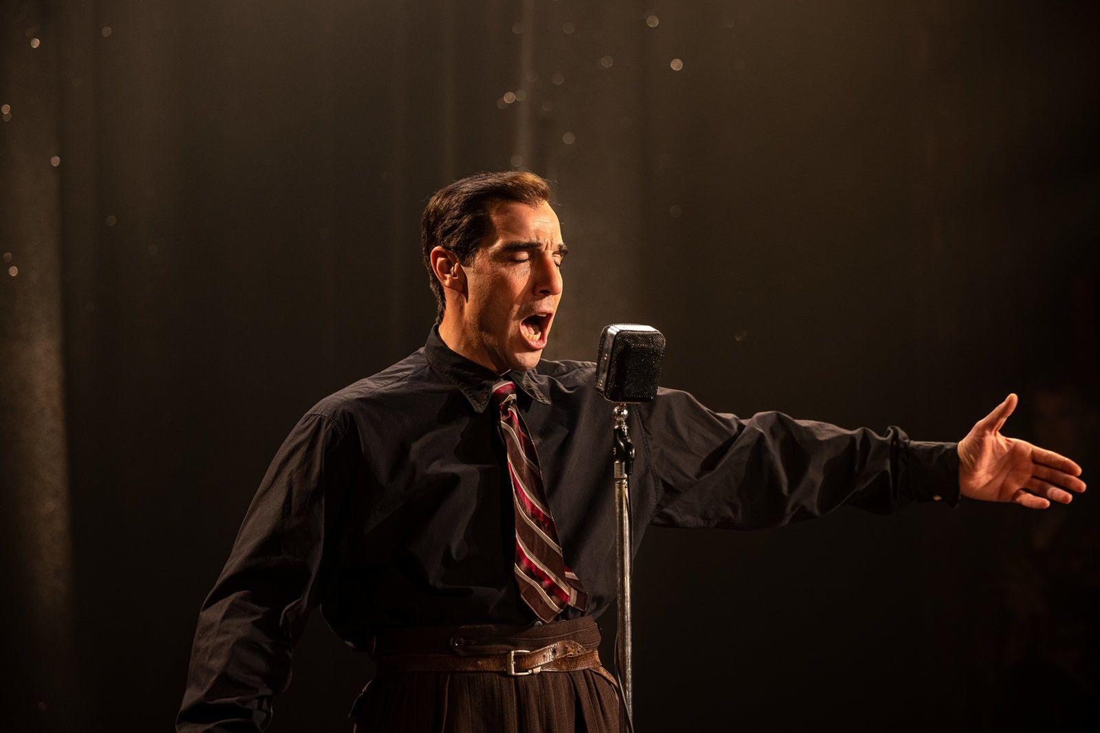 Ein Mann singt in ein Vintage-Mikrofon, trägt ein schwarzes Hemd und eine gestreifte Krawatte, mit ausgestrecktem Arm vor dunklem Hintergrund.