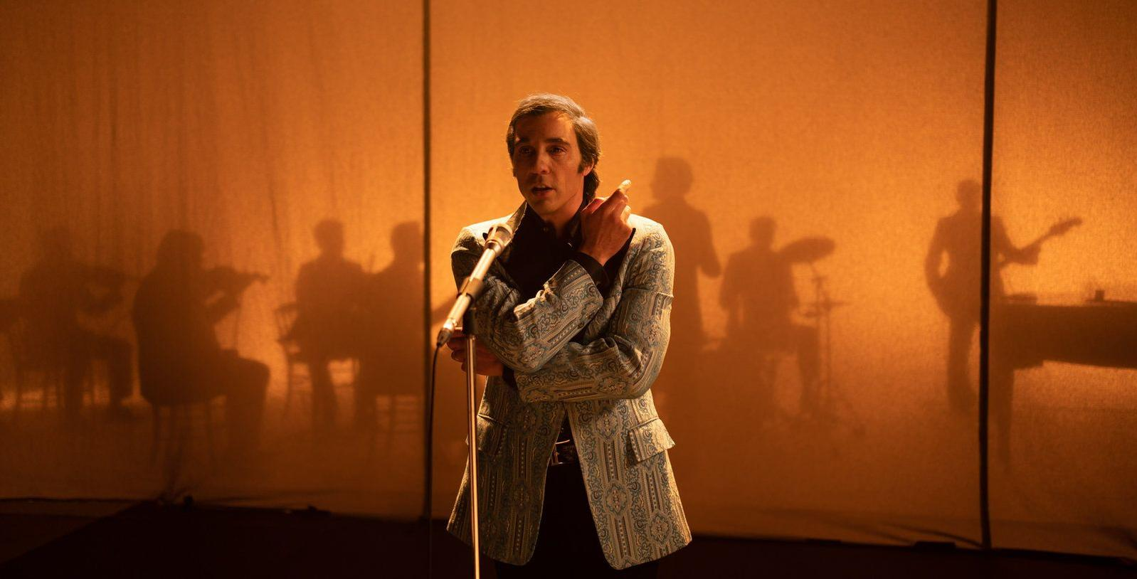 Ein Mann in einer gemusterten Jacke steht auf einer Bühne an einem Mikrofon. Im Hintergrund sind Silhouetten von Musikern vor einem orangefarbenen Hintergrund.