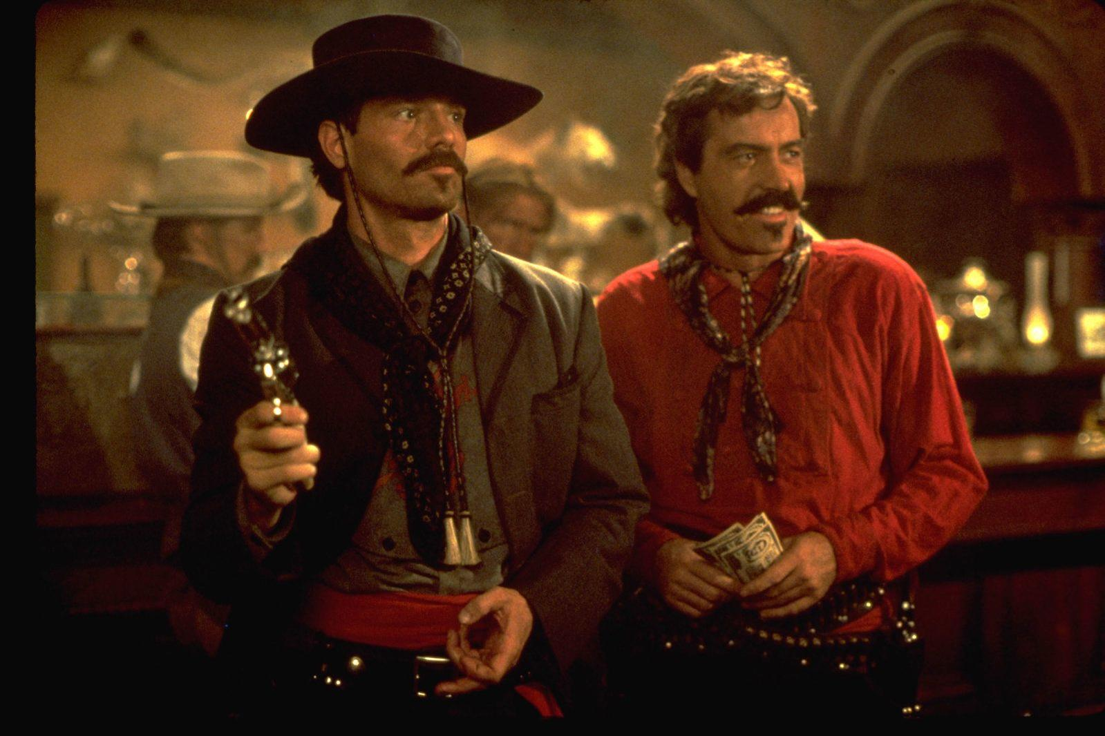 Zwei Männer im Western-Look stehen in einer Bar. Einer hält eine Waffe, der andere Geld.