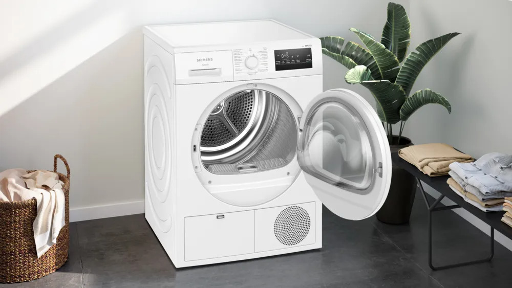 Een witte Siemens droger met een open deur. Een wasmand links en een plant rechts. Kleding op een tafel.