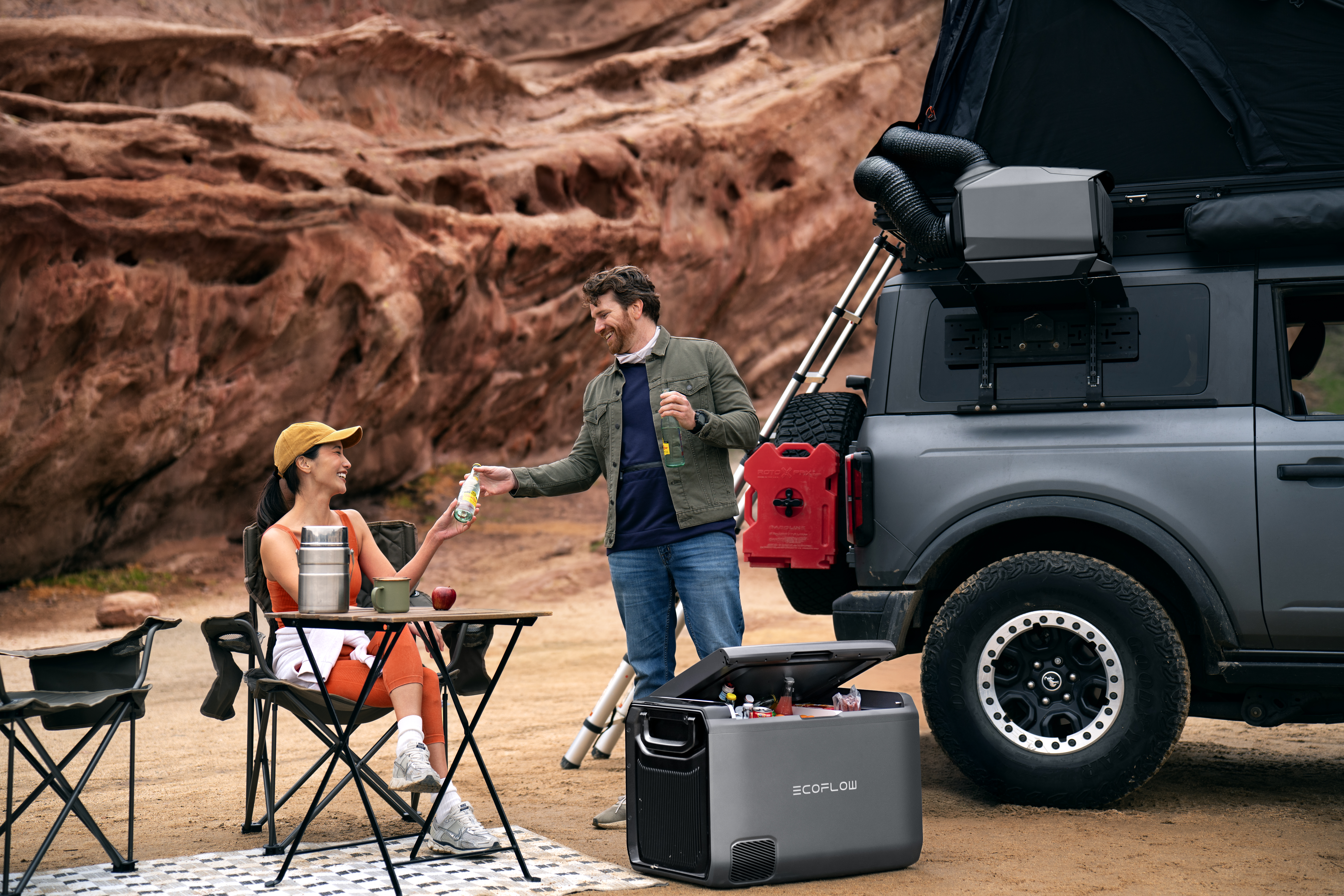 Ein Mann bietet einer Frau an einem Campingplatz ein Getränk an. Im Hintergrund steht ein Auto, und eine Kühlbox ist geöffnet. Rote Felsen und Himmel sind sichtbar.