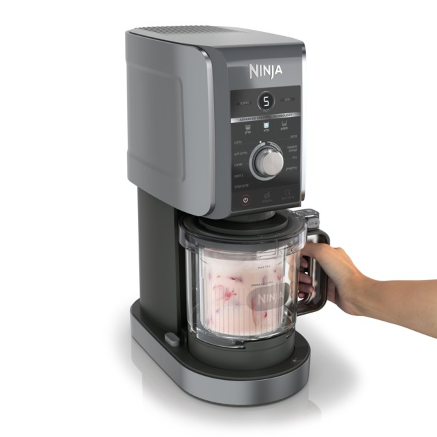 A Ninja Creami appliance. A hand holds the handle. The container has pink ice cream with visible strawberry pieces.