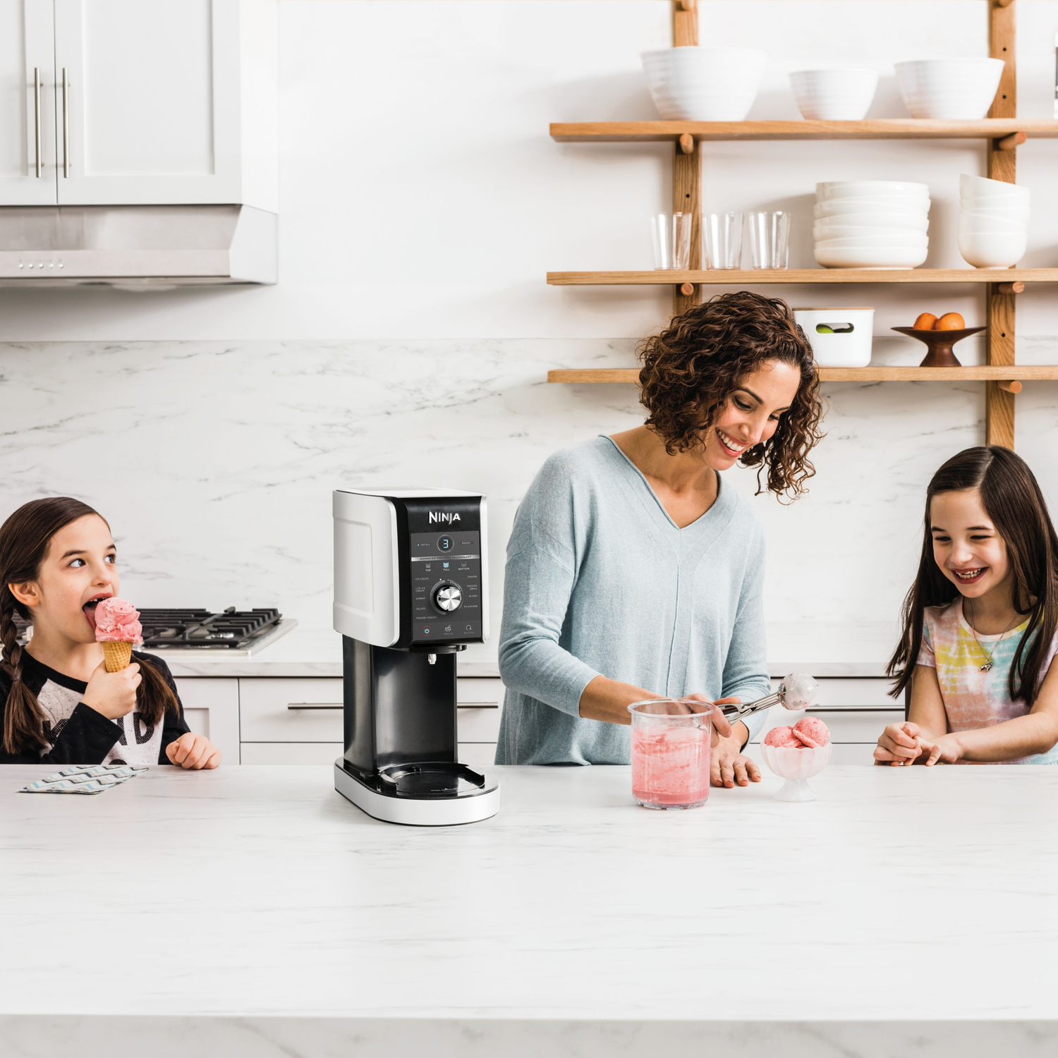 A woman serves ice cream to two girls. One girl eats ice cream in a cone. A machine is on the counter.