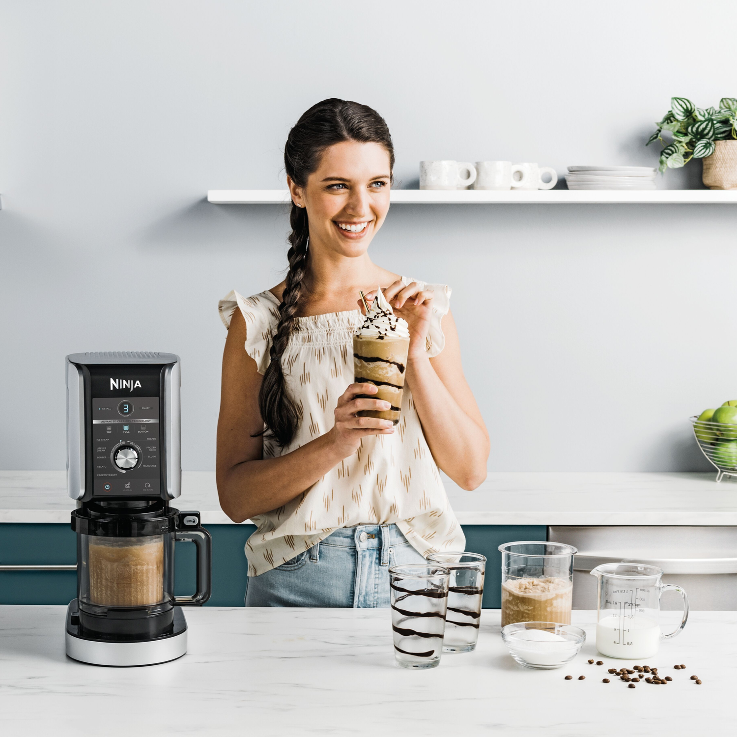Woman holding a milkshake with blender in the background. Ingredients like milk, sugar, and coffee beans are on the counter.