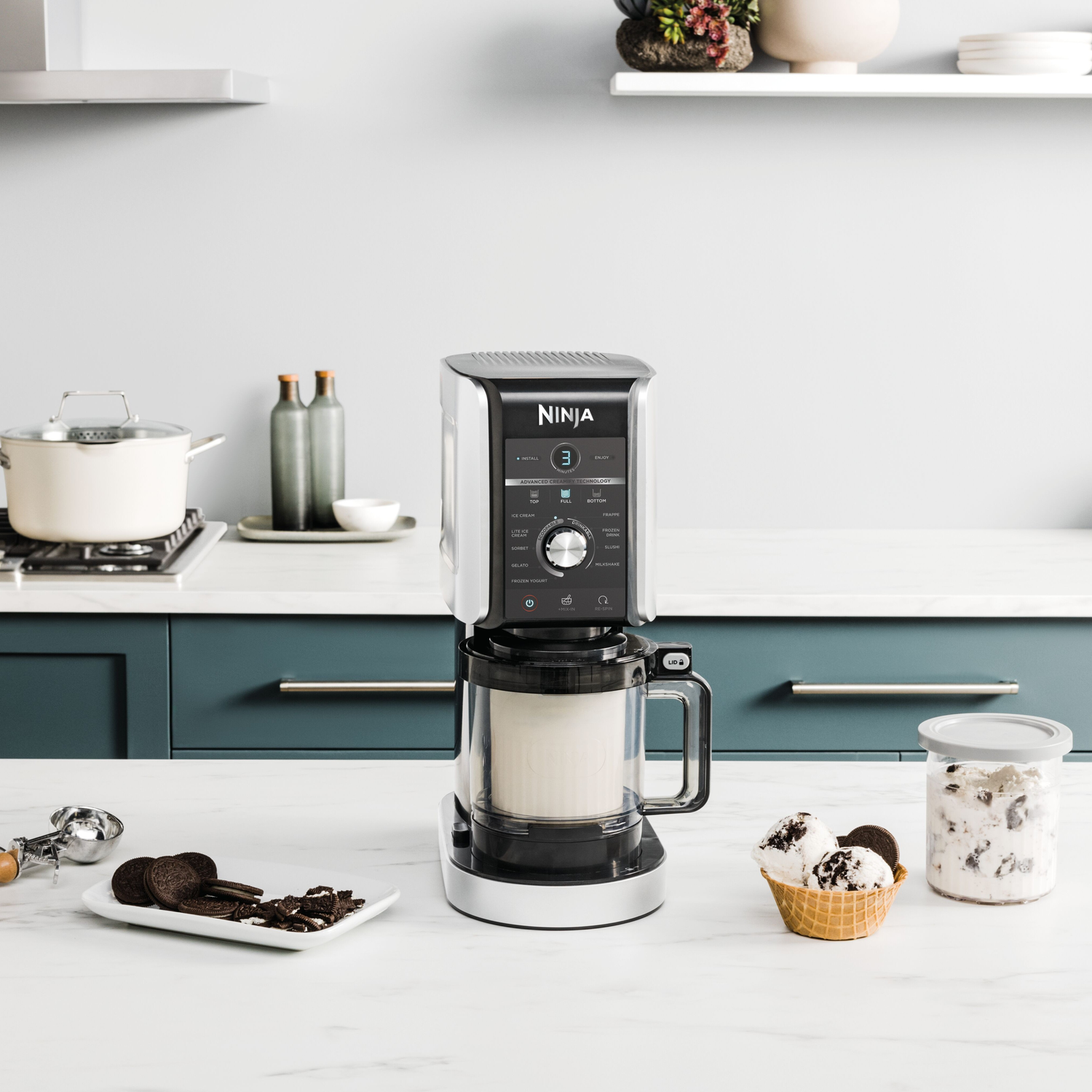 Ninja CREAMi machine. Oreo cookies on a plate, ice cream in a waffle cup, and a jar of ice cream. Kitchen background.