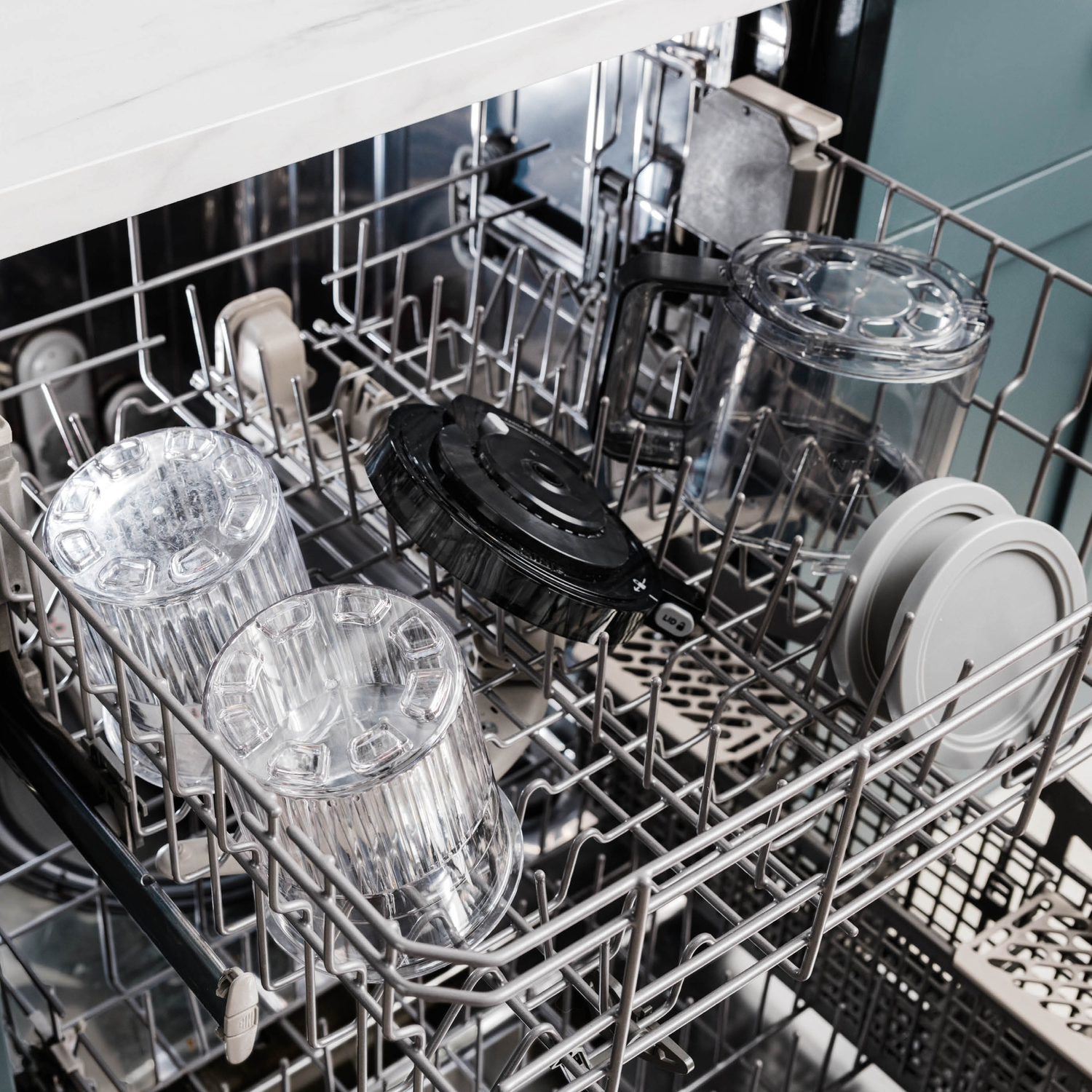 Dishwasher interior loaded with clear plastic cups and lids. A black container with a lid and other items are also present.